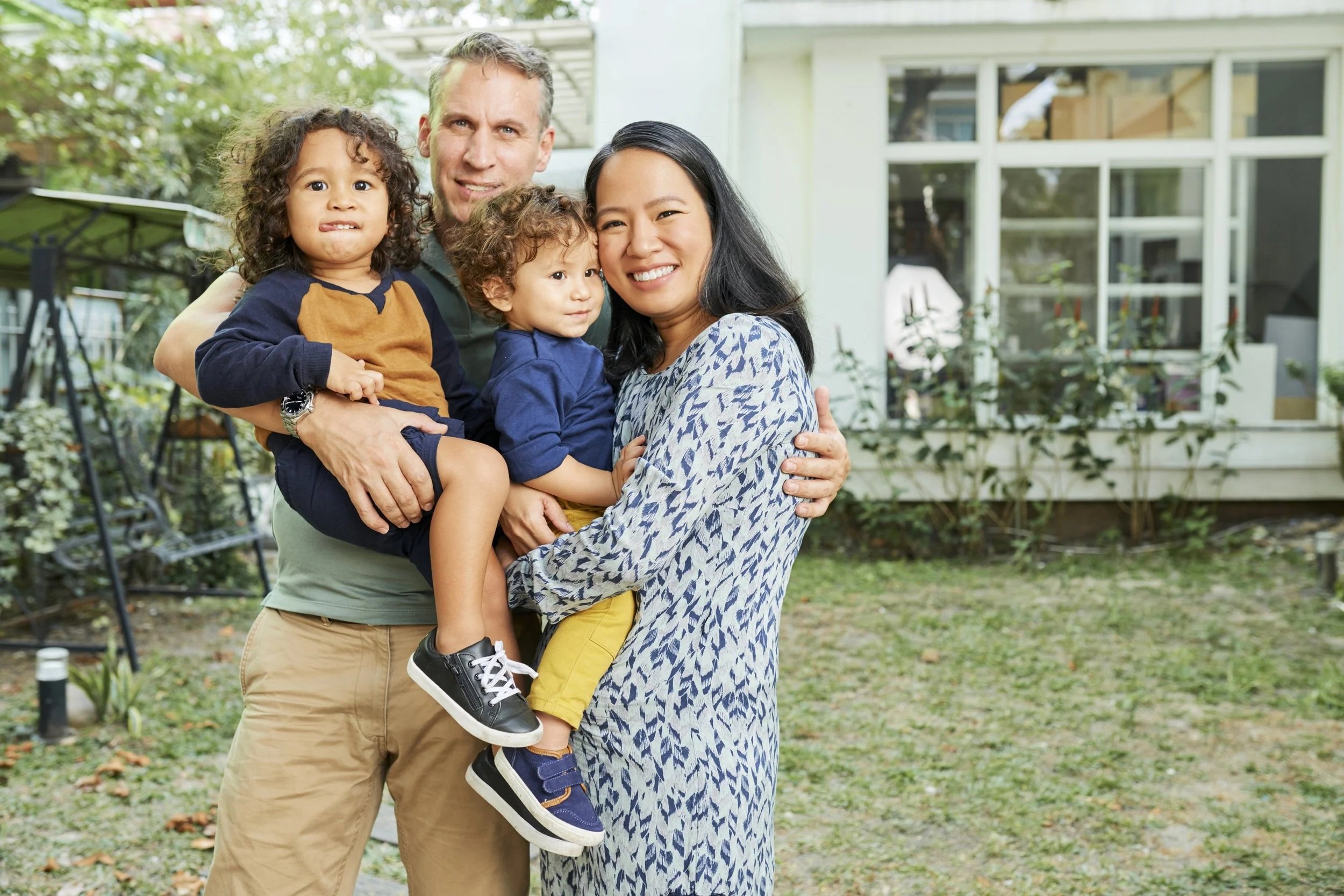 A diverse family of three adults and two children smiling and embracing outside in a front of house. The children have a better connection with their parents because of speech therapy for kids in Maui, HI.