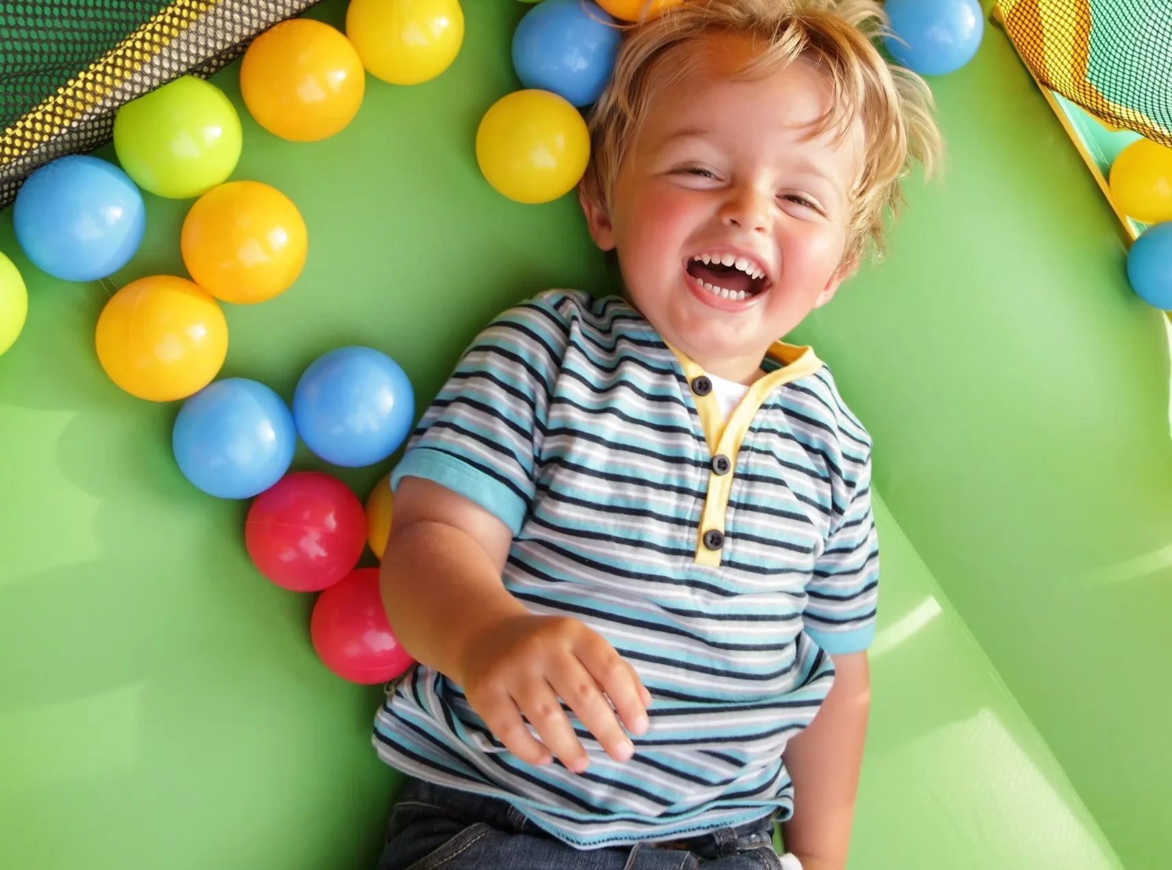 A young boy with blonde hair lying in a ball pit filled with colorful plastic balls, laughing and smiling during occupational therapy for kids in Maui, HI.