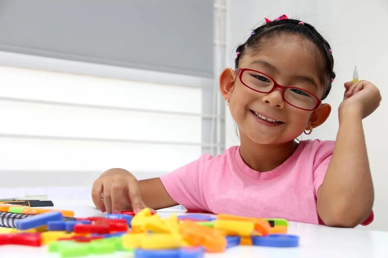A young girl with glasses and a pink shirt smiling while playing with colorful game pieces during occupational therapy for children in Maui, HI.