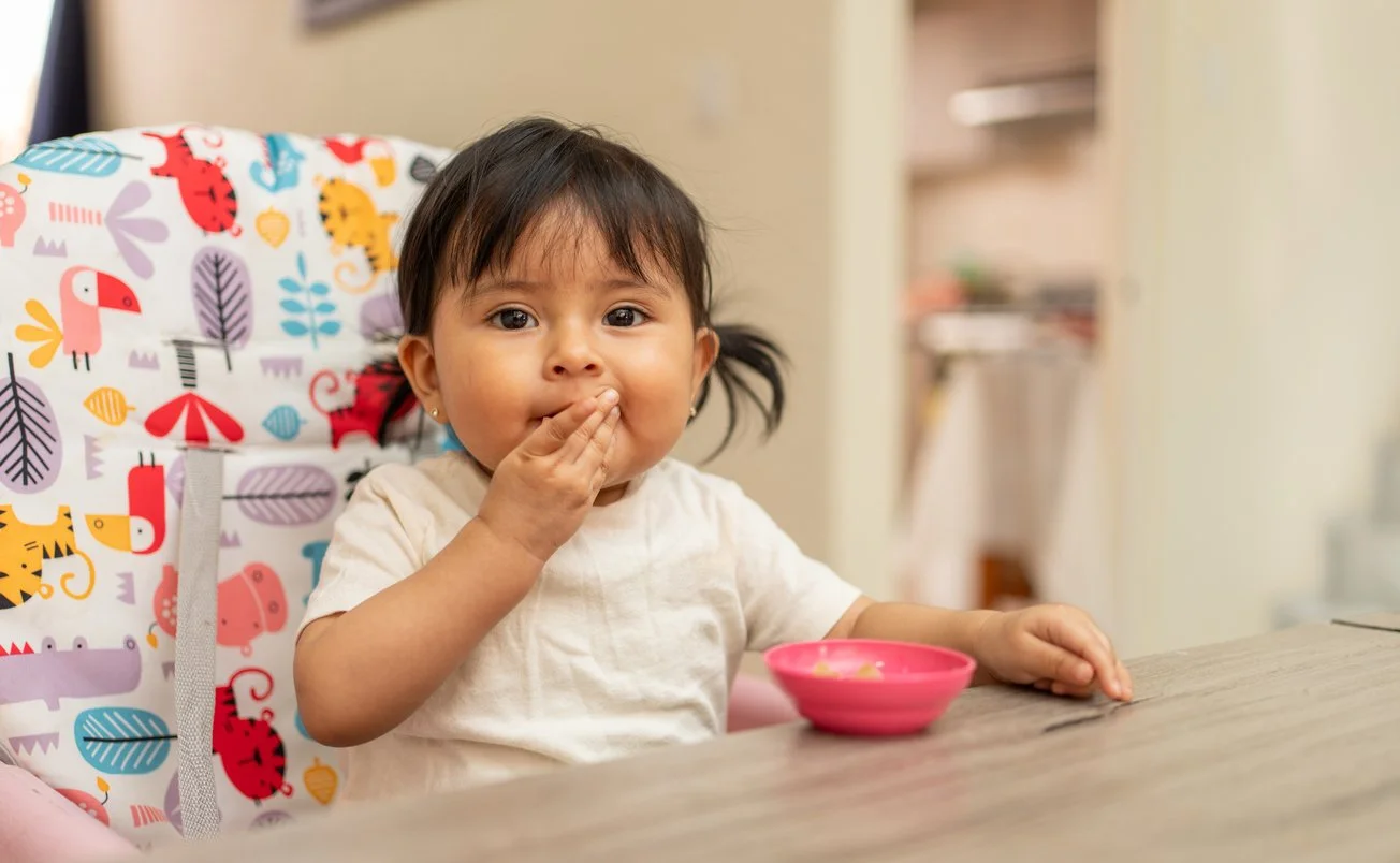 Young girl sitting in a highchair, eating from a pink bowl on a wooden table during feeding therapy for kids in Maui, HI.
