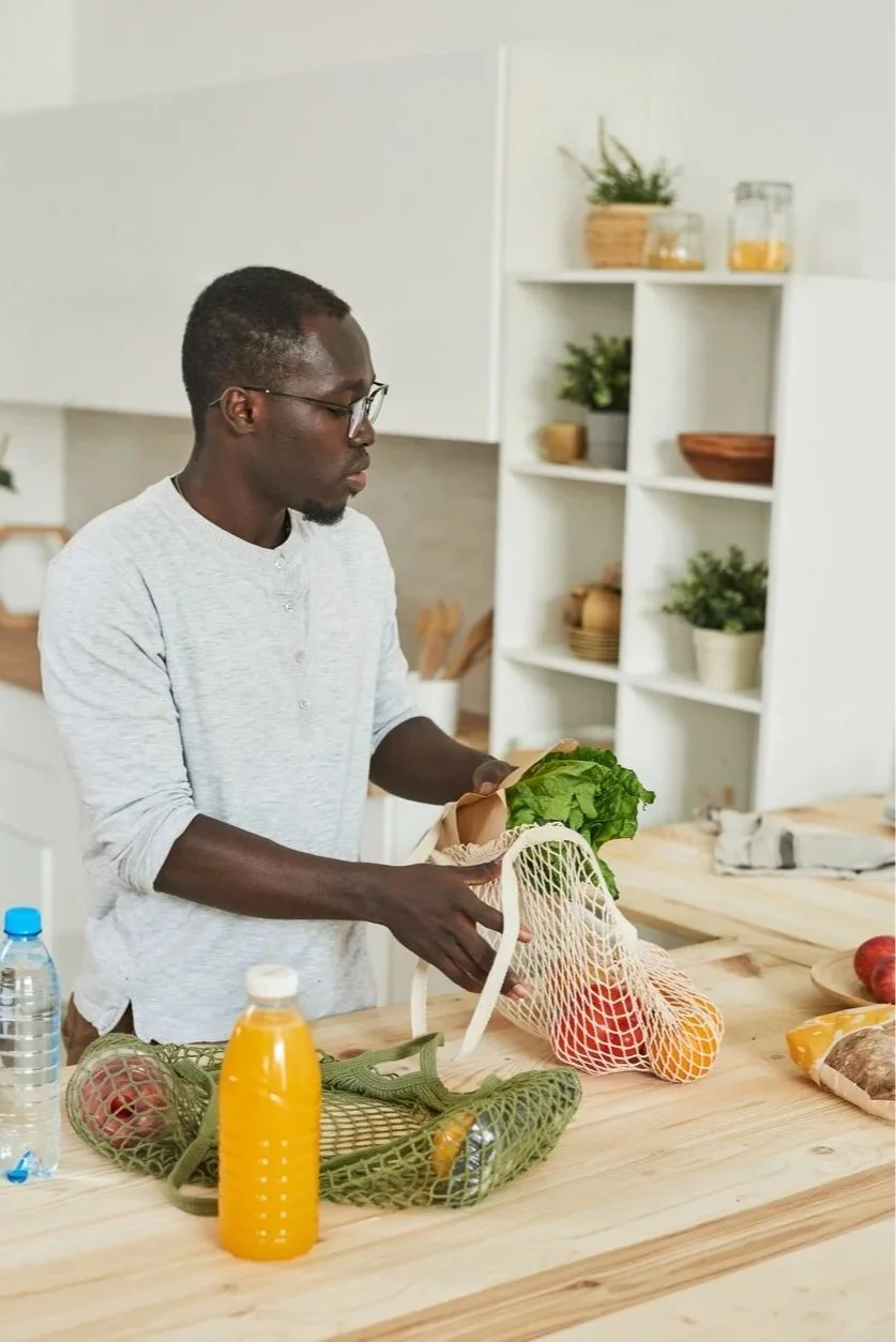 A man with improved life skills and daily living after occupational therapy for adults in Maui, HI, packing fresh vegetables into a reusable shopping bag in a kitchen with white shelves and potted plants.