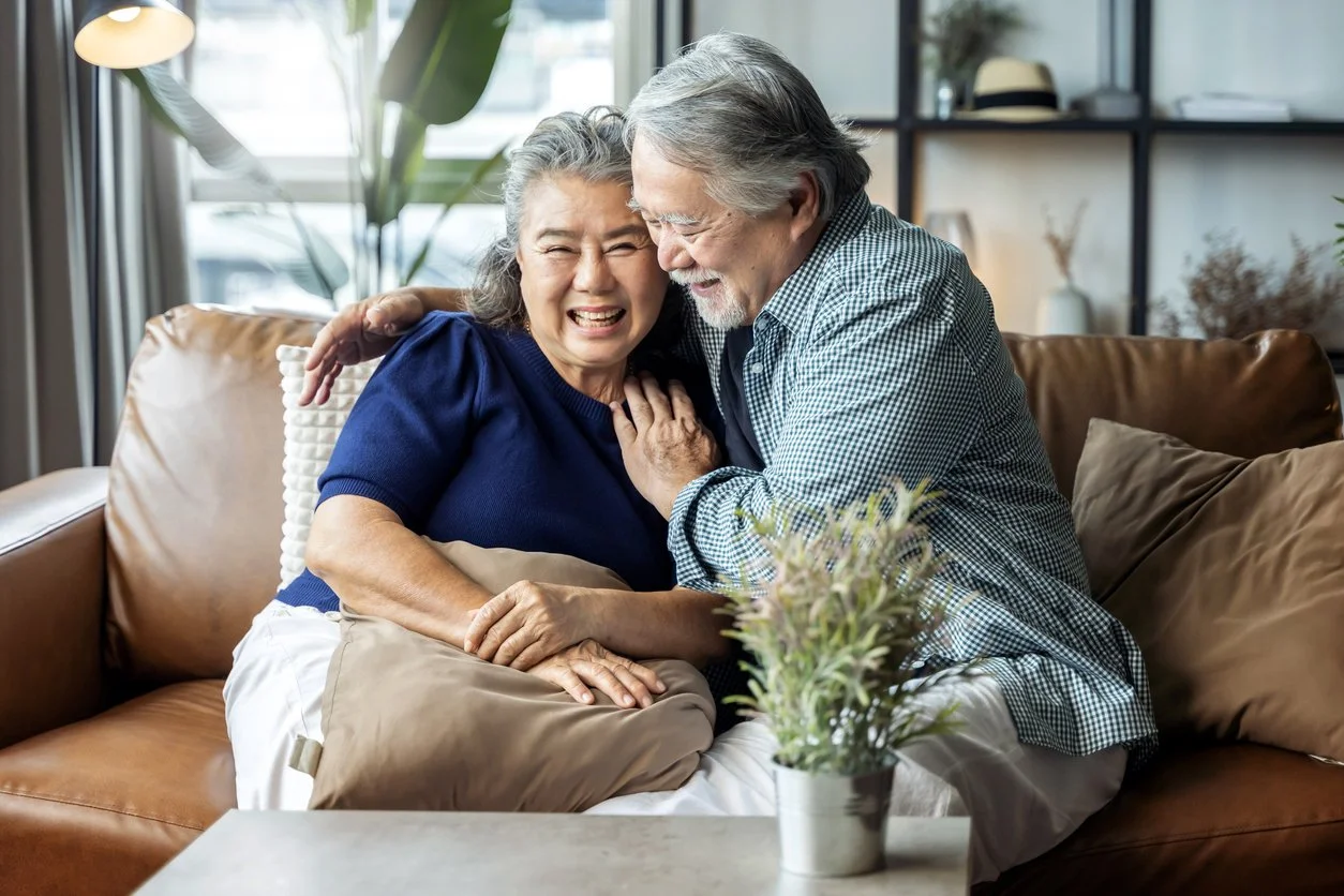 An elderly woman and man sitting on a brown leather sofa, smiling and embracing each other, with the woman having improved independence and cognitive skills after speech therapy and occupational therapy for adults in Maui, HI.