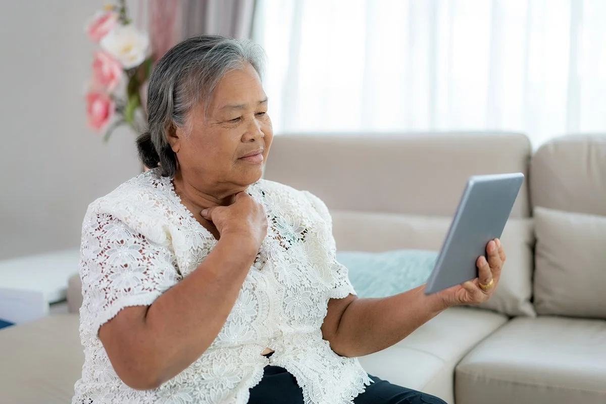 An elderly woman with gray hair sitting on a beige sofa in a bright living room, looking at a tablet while receiving online voice therapy in Maui, HI.