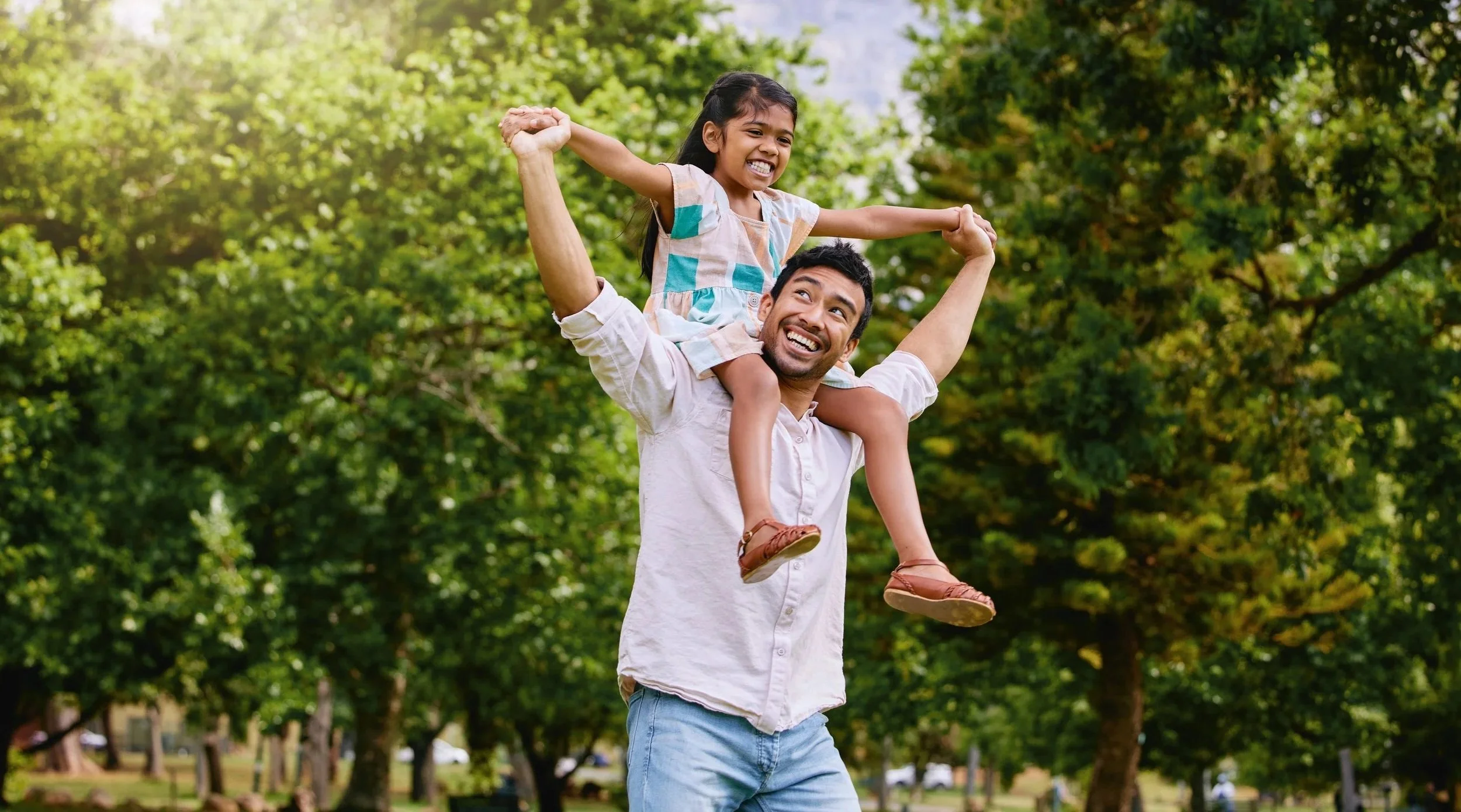 A man holding a young girl on his shoulders in a park with green trees, both smiling and enjoying a sunny day, communicating with less frustration because of speech therapy for kids in Maui, HI.