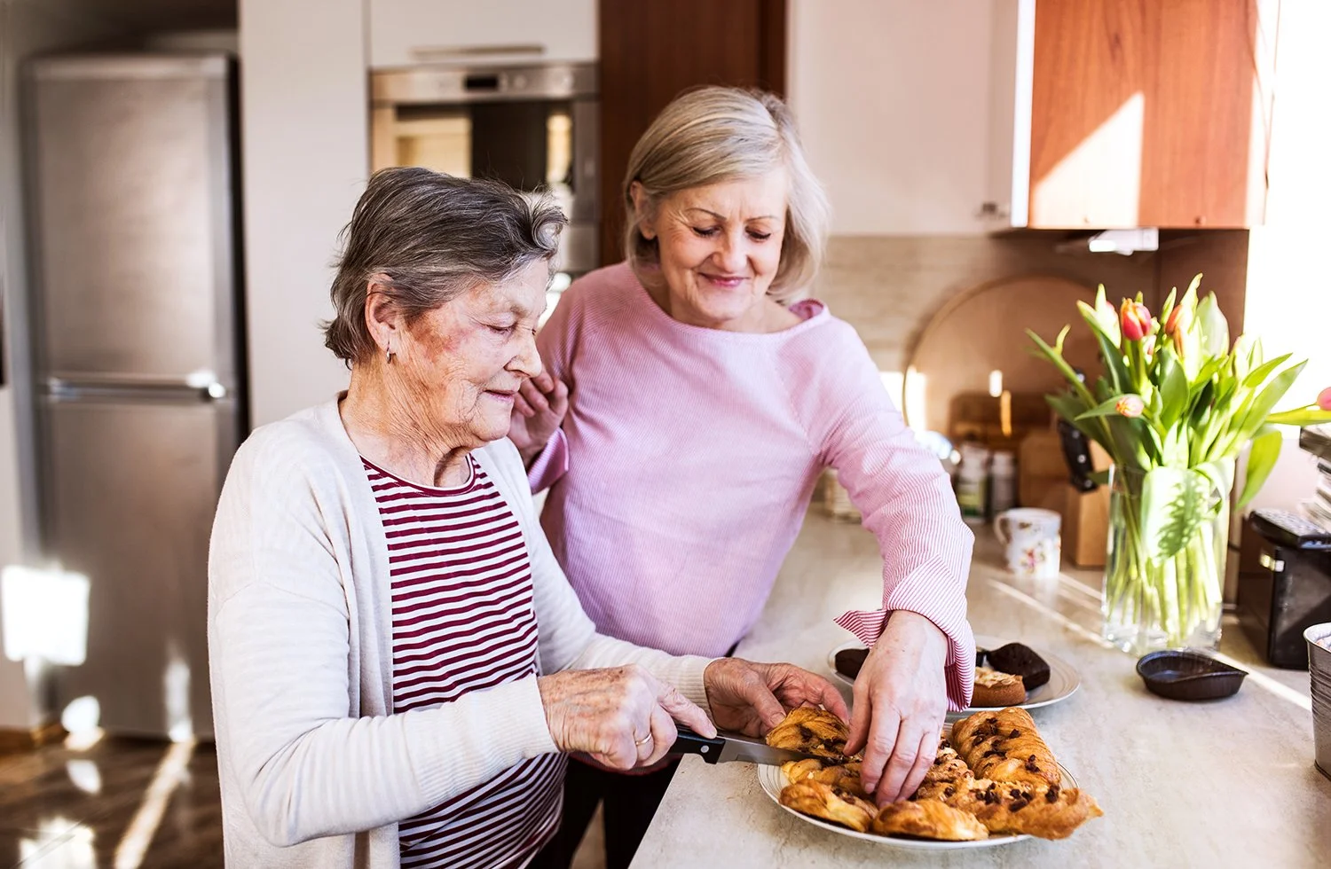An elderly woman able to enjoy mealtimes again because of swallow therapy in Maui, HI, cutting a plate of pastries in a kitchen with sunlight and a vase of pink tulips on the counter.