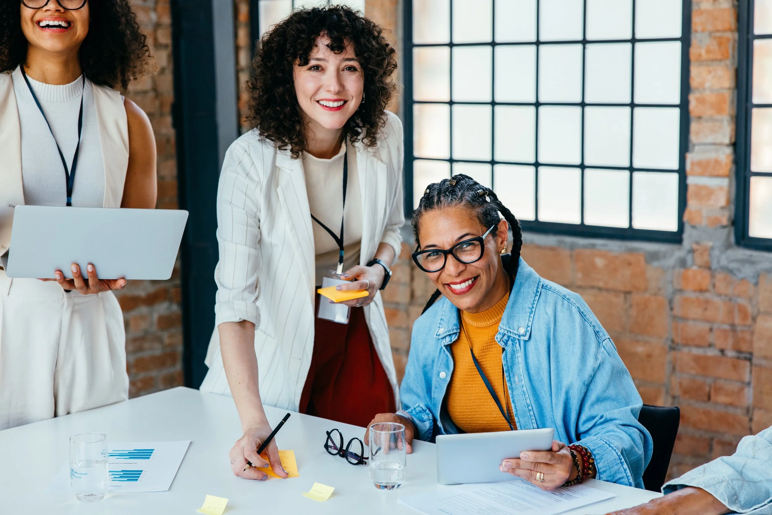 LGN Collab: Four women in a meeting room, smiling and engaging with each other. One woman is seated with a tablet, another is standing with a notepad, and two women are holding a laptop and a notepad. The room has brick walls and large windows.