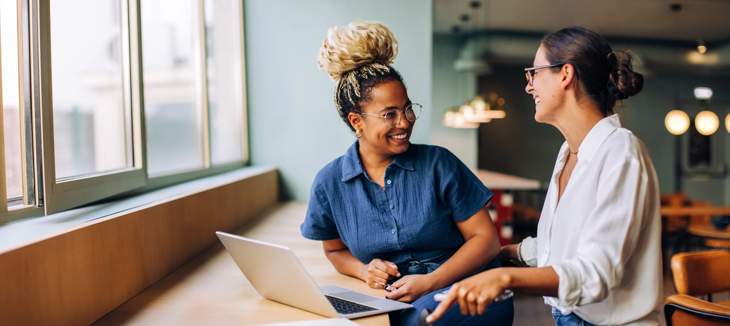 LGN Collab: Two women happily talking about their team operations in a workspace near a window, with a laptop open on the table between them.