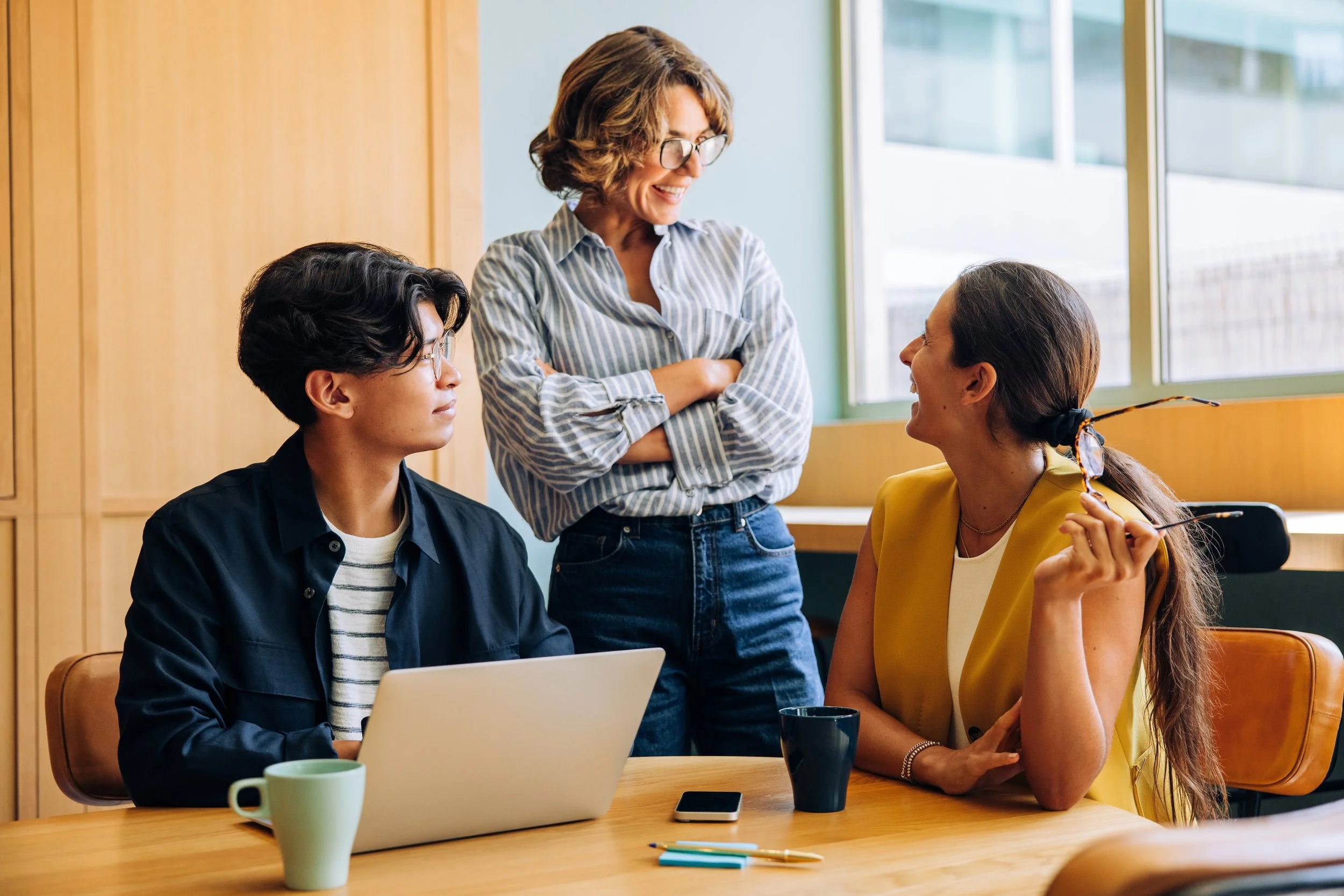 LGN Collab : Three women having a conversation in an office, with two sitting at a table with a laptop and coffee mugs, and one standing smiling.