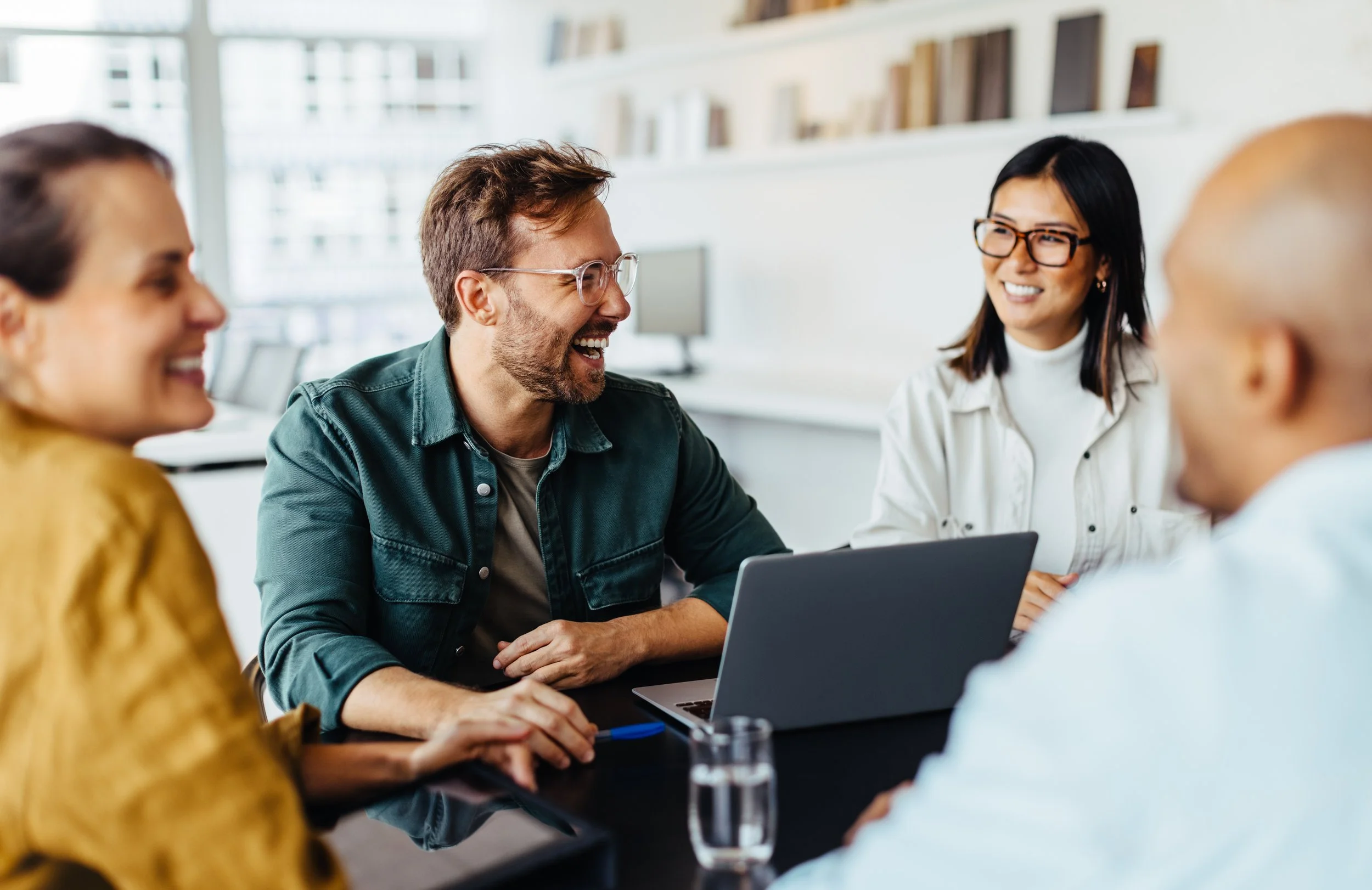 LGN Collab: A group of four diverse people sitting around a table in an office, smiling and engaging in conversation, with a laptop and glass of water on the table.