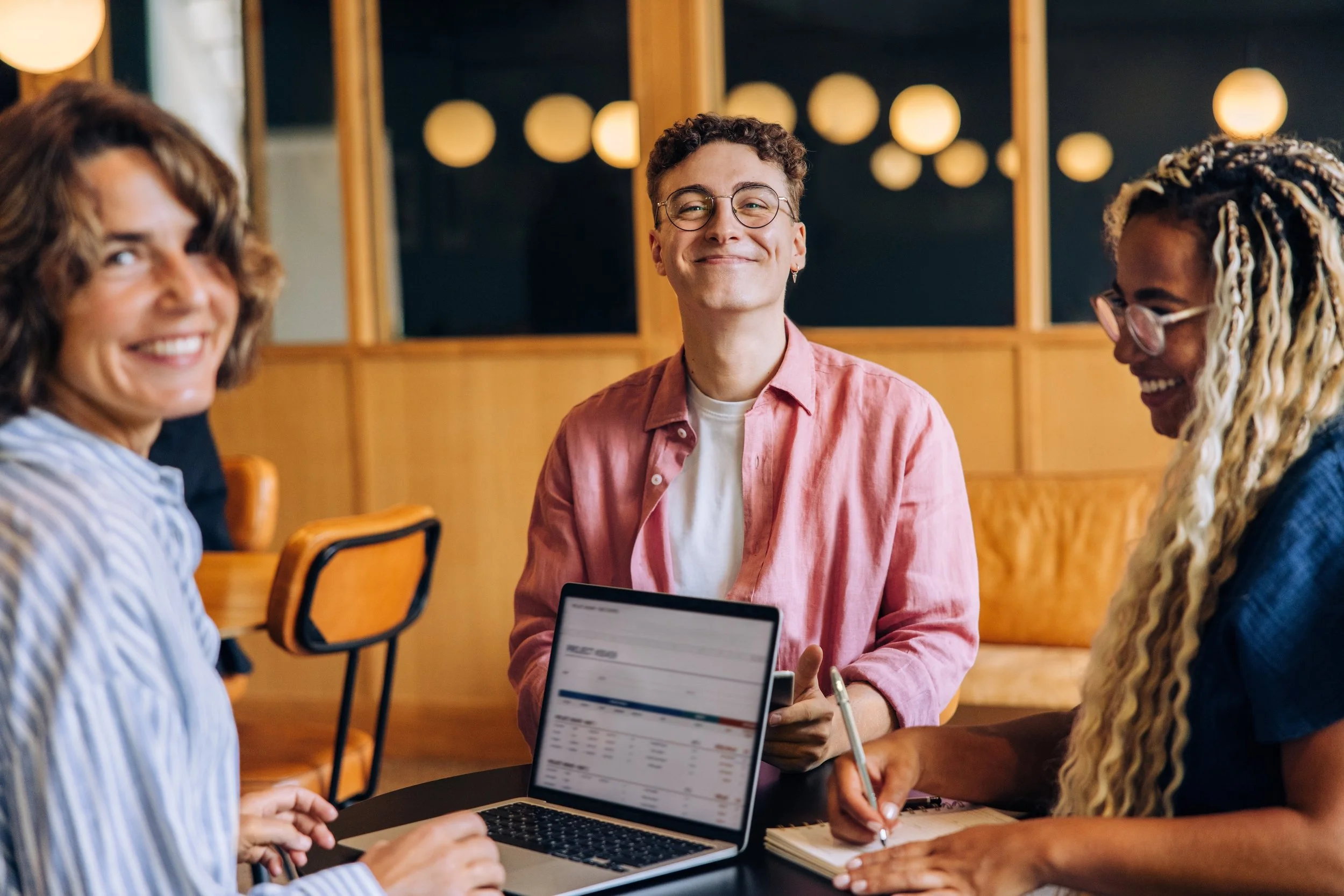 LGN Collab: Three people smiling, gathered around a table with a laptop and a notepad, in a cozy room with wooden walls and warm lighting.