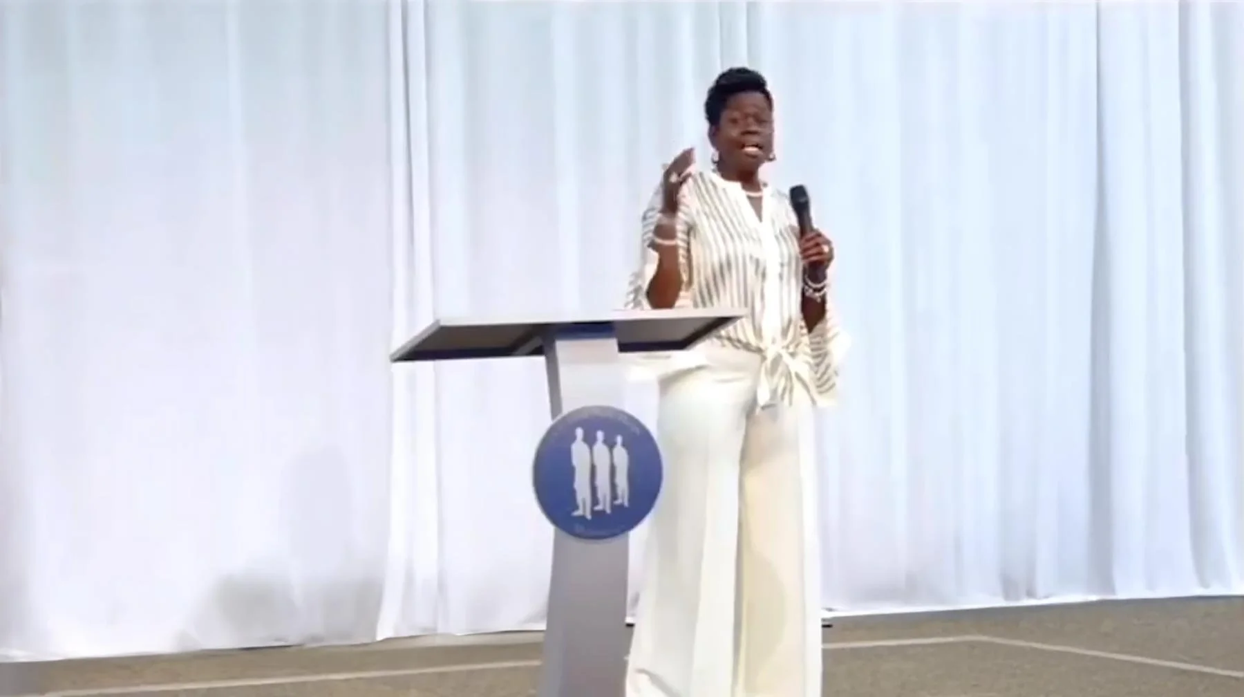 Joanne Mitchell-McLaren speaking into a microphone on stage, standing behind a podium with a symbol of three figures. She is wearing a striped blouse and white pants, with a white curtain background.