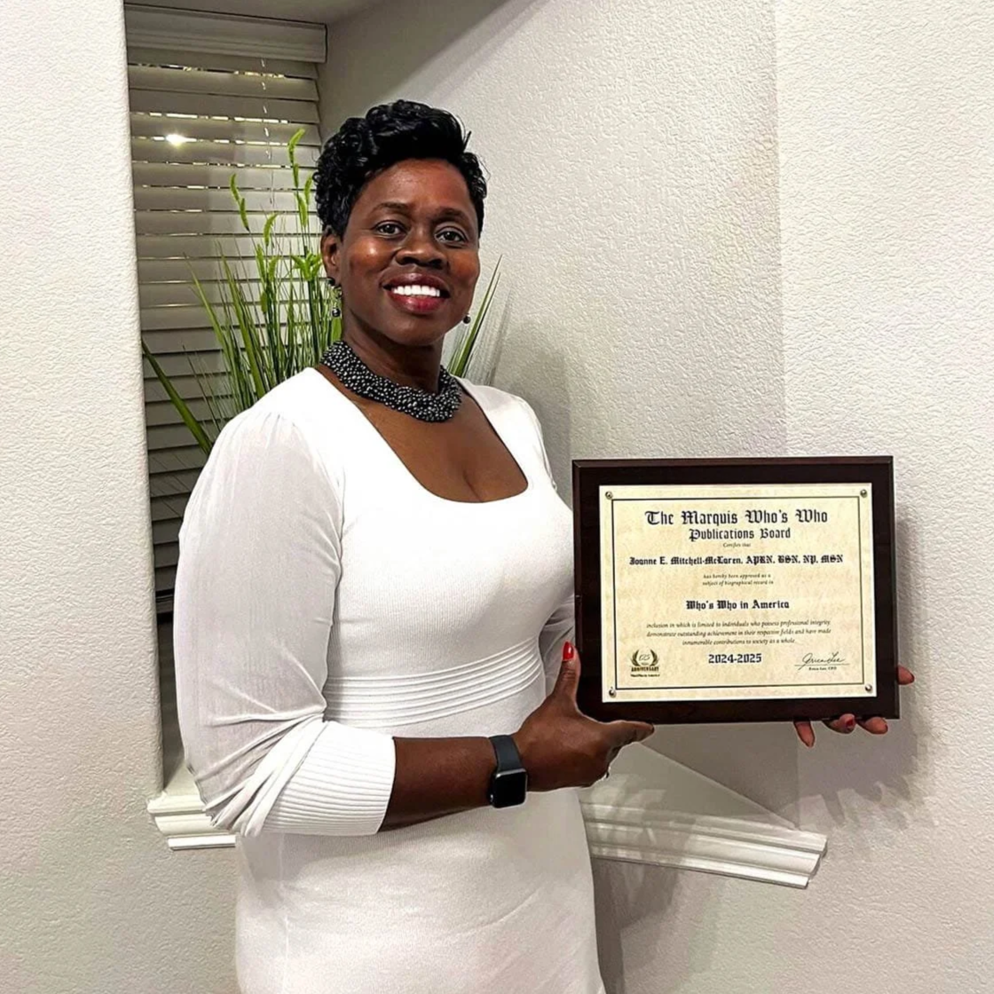 Joanne Mitchell-McLaren in a white dress is smiling and holding a framed certificate, standing inside a room with a plant and window blinds in the background.