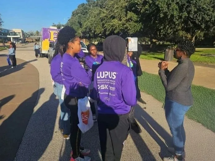 Group of people, mostly women, gathered outdoors in a park, wearing purple shirts with 'Lupus Foundation of America' on the back, listening to Joanne Mitchell-McLaren speaking.