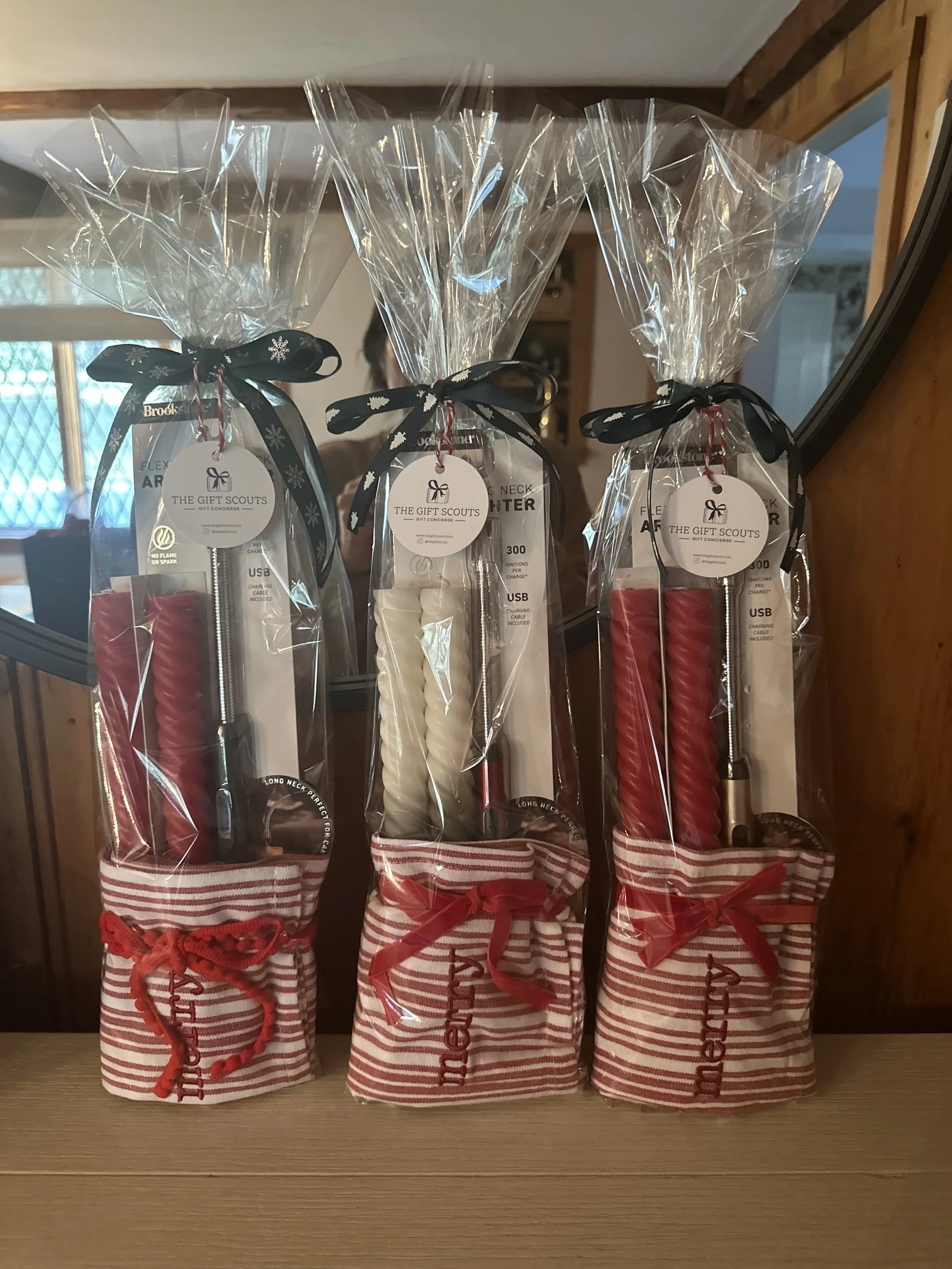 Three gift packs containing twisted candles and USB charging cables, wrapped in clear plastic and tied with black ribbons, sitting on a wooden surface.
