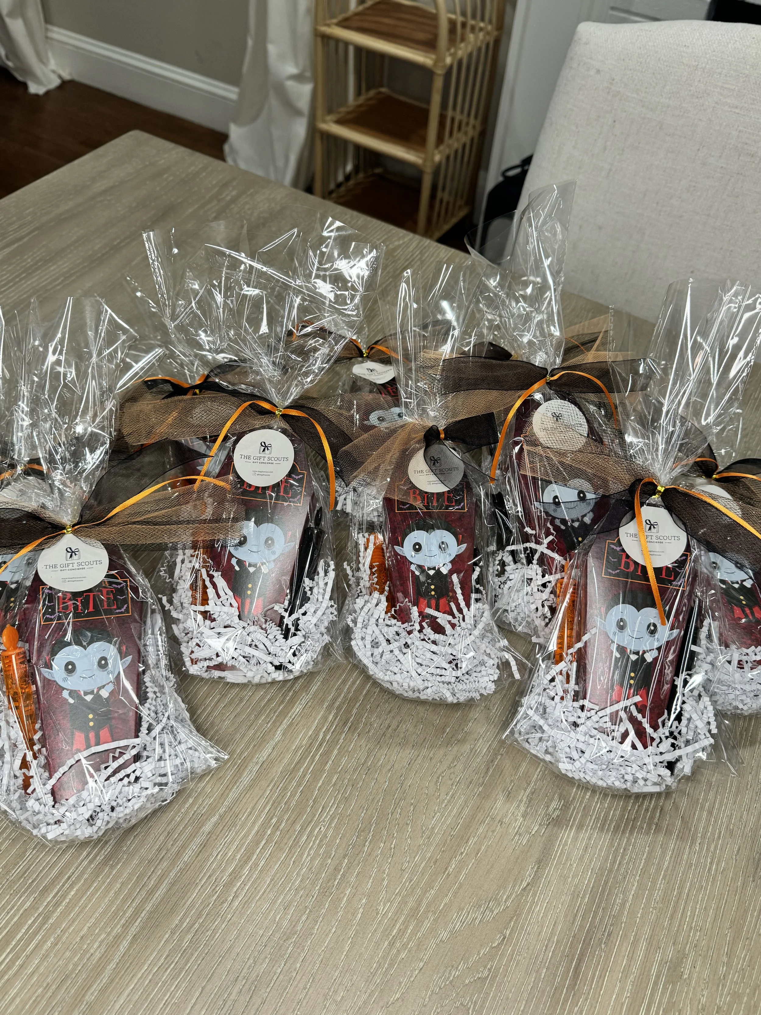 Packaged Halloween-themed treat bags on a wooden table, featuring a cartoon vampire. The bags are clear, with black and orange ribbons and shredded paper inside.