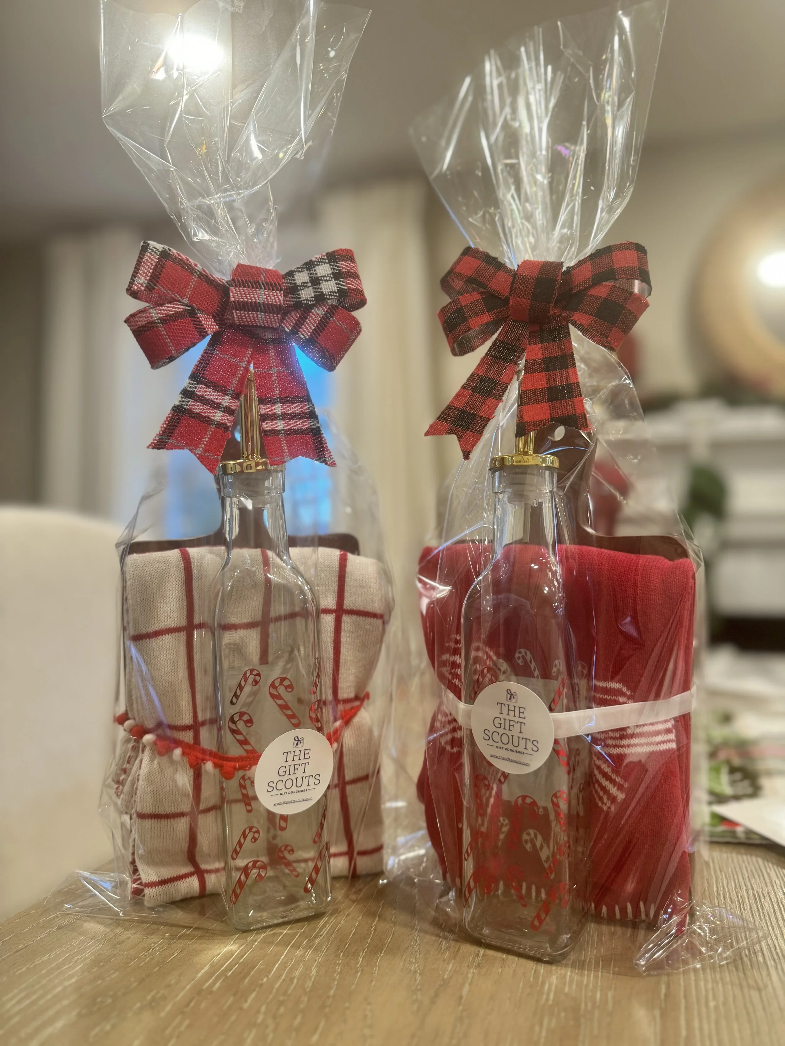 Gift sets containing a bottle with a plaid fabric sleeve, wrapped in clear plastic with a red and black plaid bow and a candy cane design sticker, on a wooden table.