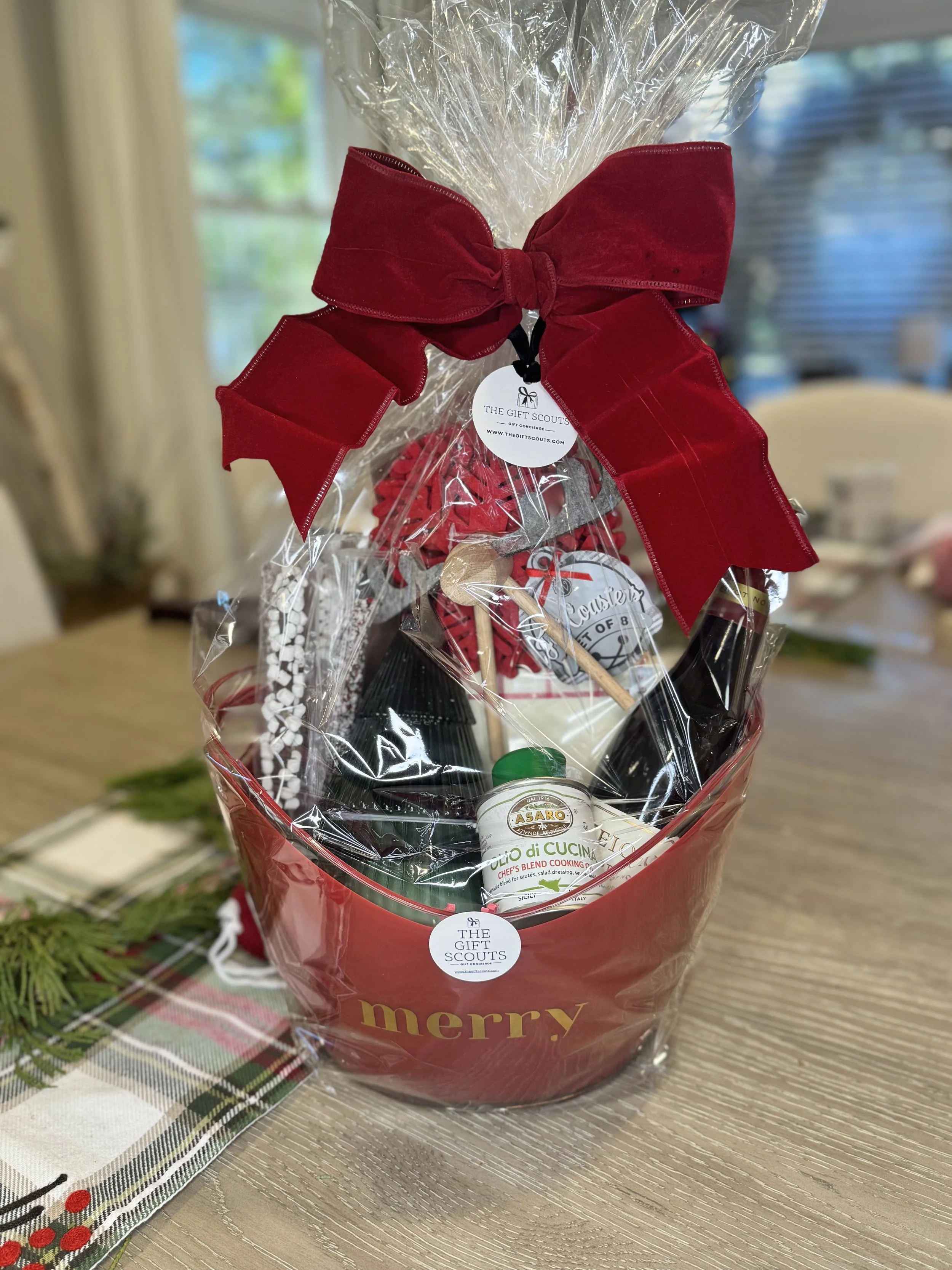 Gift basket wrapped in clear plastic with a large red bow, containing food and cooking items, with 'merry' written in gold on the basket.
