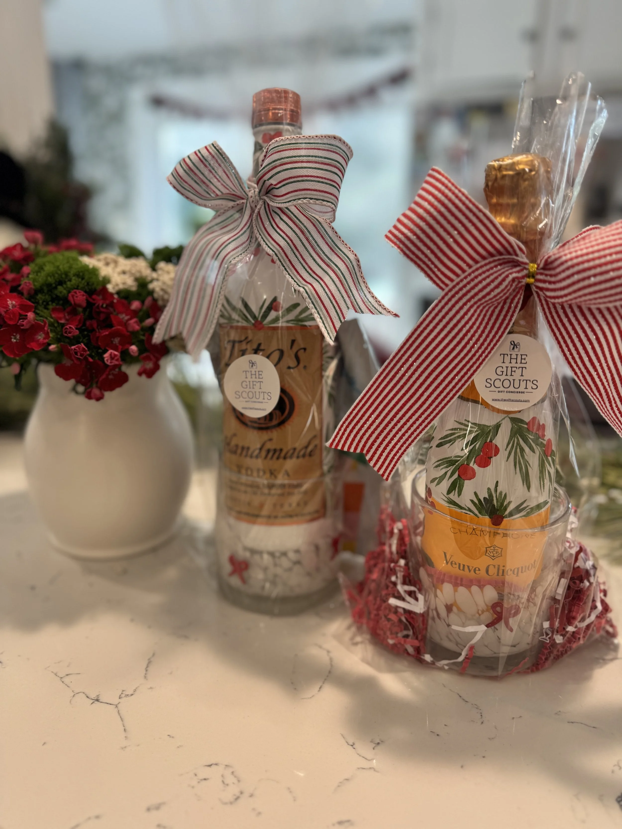 Gift bottles wrapped in decorative ribbons and tissue paper, with indoor Christmas decorations and potted flowers in the background.