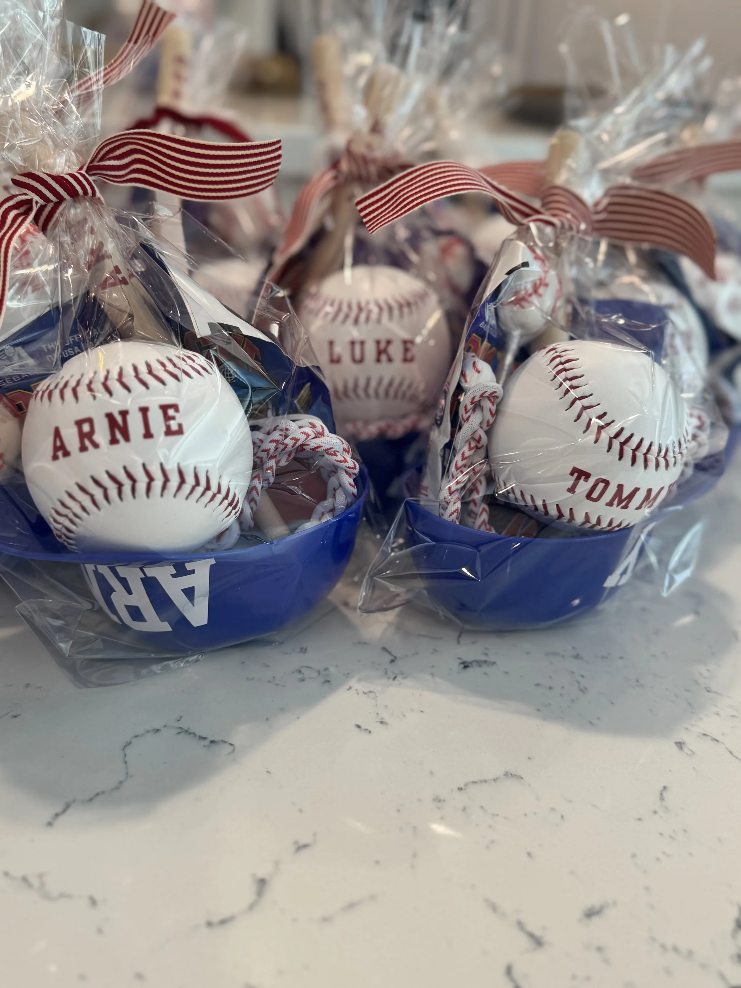 Gift baskets containing baseball-themed items, wrapped in plastic, with red and white striped ribbons, labeled with names Arnie, Luke, and Tommy, on a white marble countertop.
