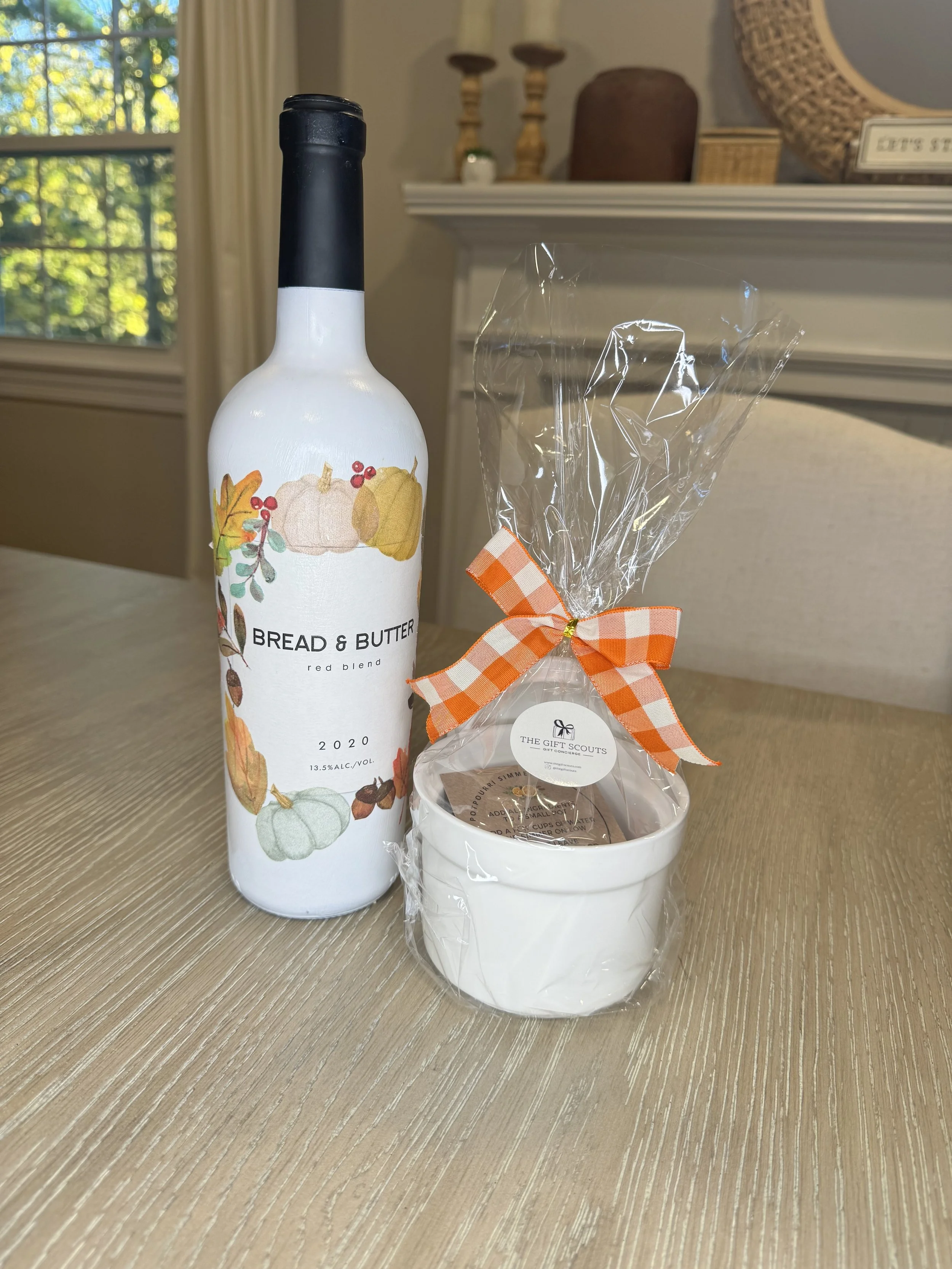 A bottle of Bread & Butter red blend wine, a small white bowl with a gift tag, and a wrapped item with an orange checkered ribbon on a wooden table. In the background, there is a fireplace mantel with candles and decorative items.