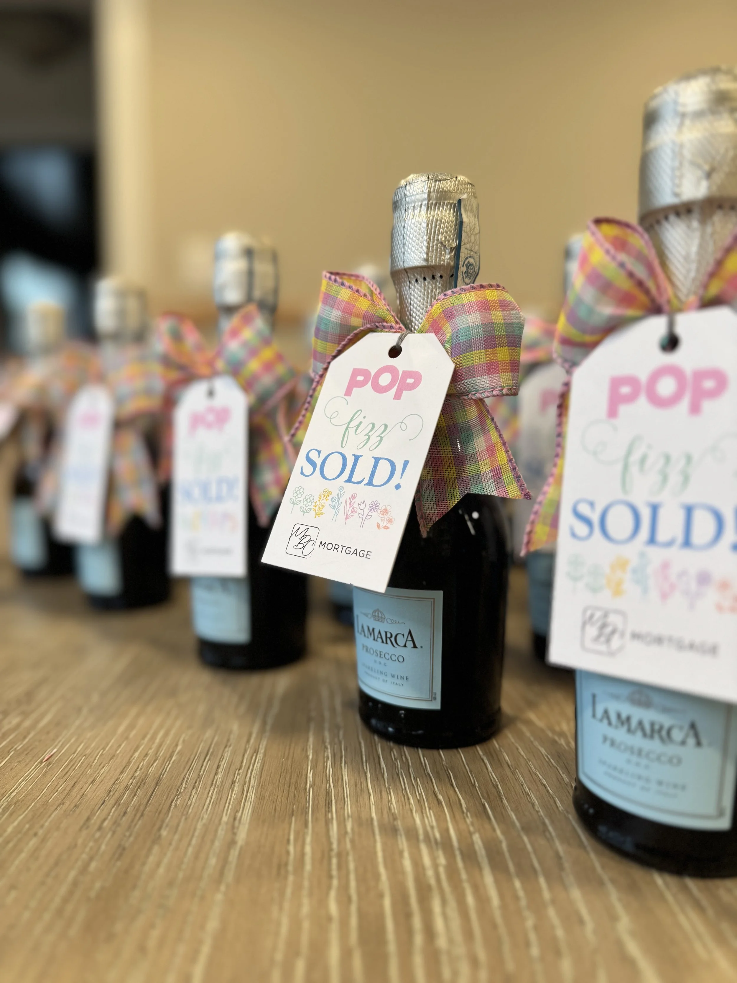 Several bottles of Lamarca Prosecco with checkered pink and yellow ribbon bows and tags that say 'POP fizz SOLD!' on a wooden surface.
