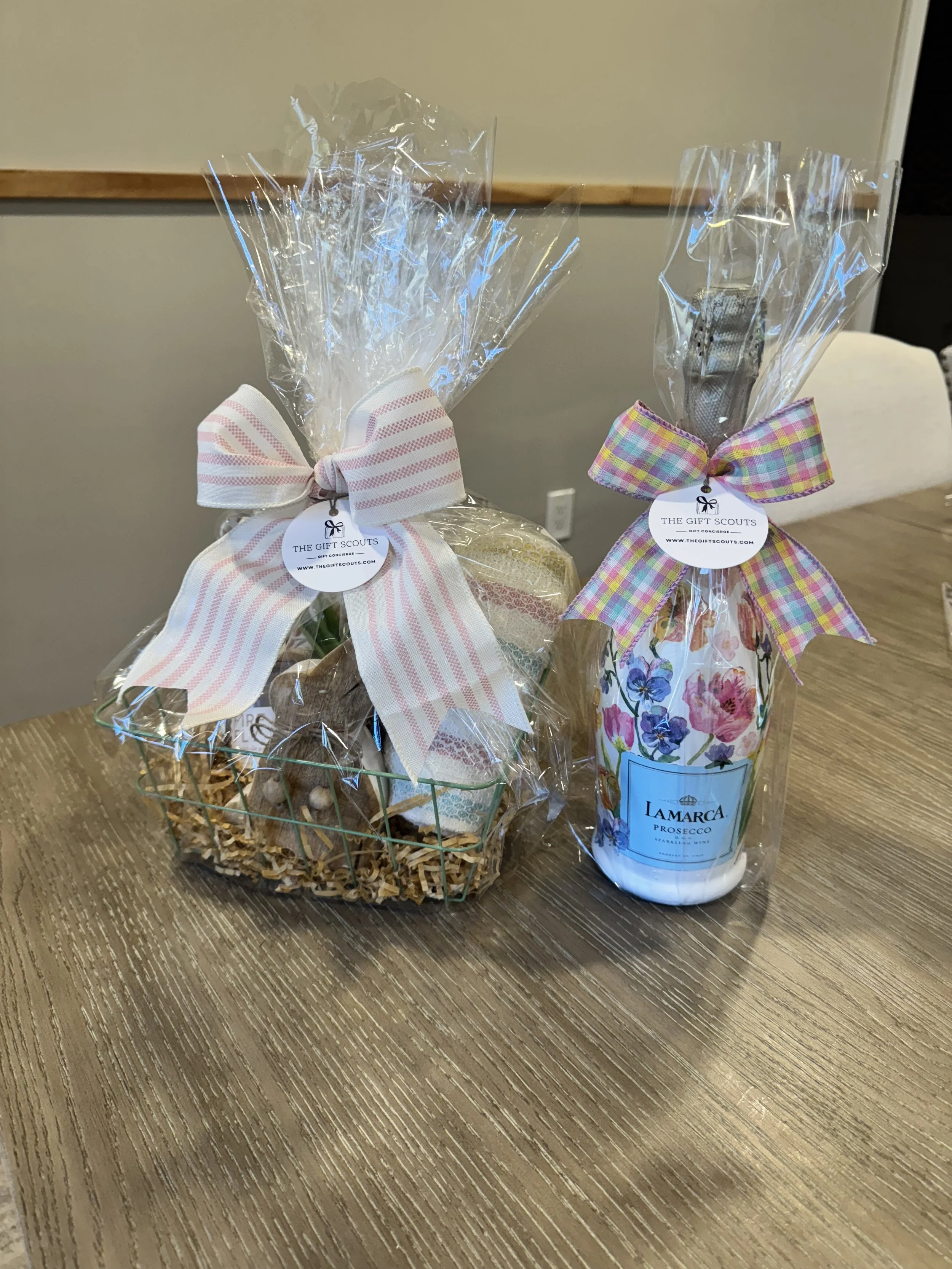 Gift basket with assorted items wrapped in clear cellophane and decorated with a pink and white striped ribbon, and a bottle of Lamarca Prosecco wrapped with a multicolored checkered ribbon.