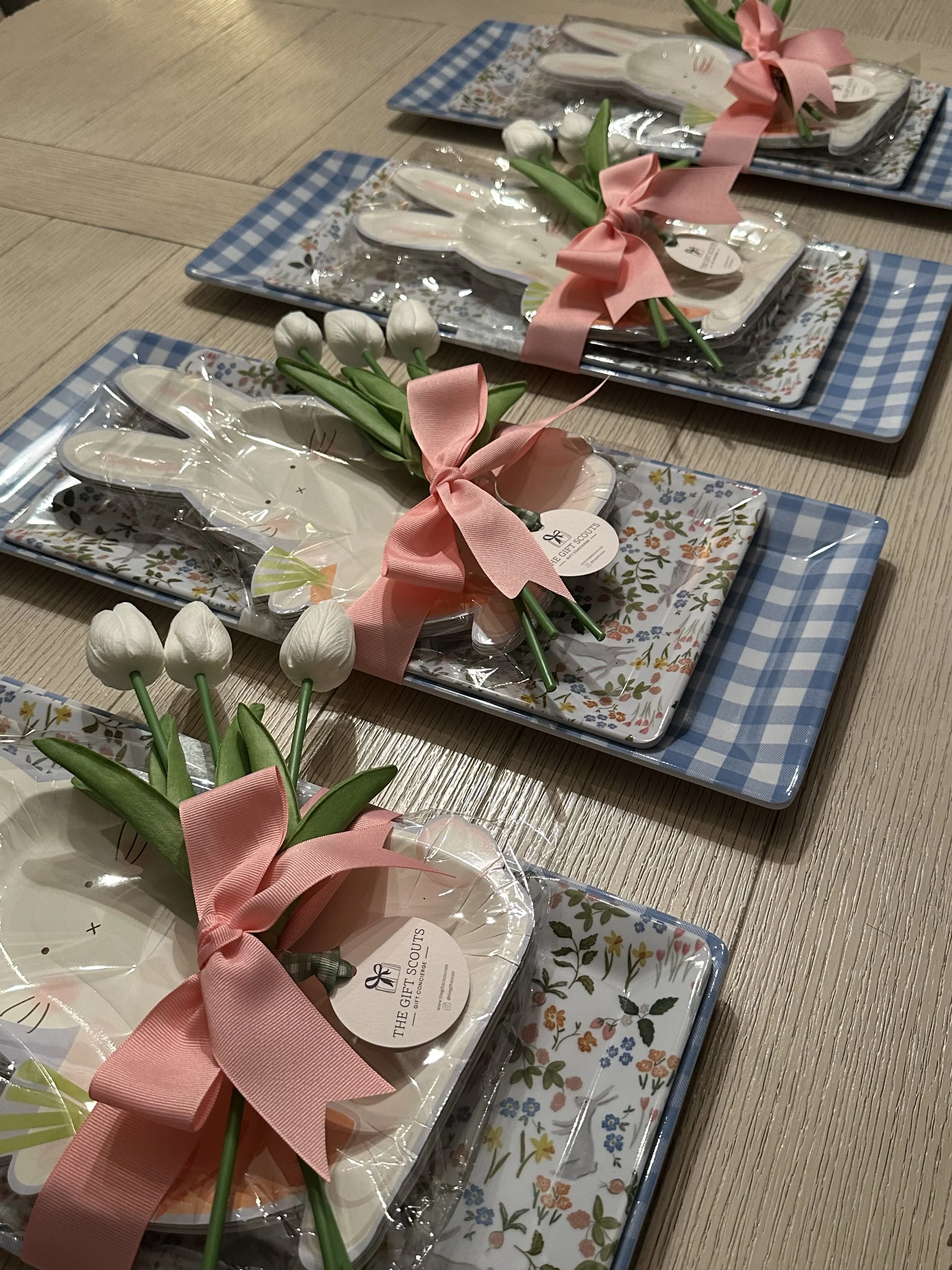 Four Easter-themed gift sets on a wooden table, each featuring a pair of white bunny-shaped utensils, pink ribbons, and small white tulip flowers, all in decorative packaging with floral and checkered patterns.