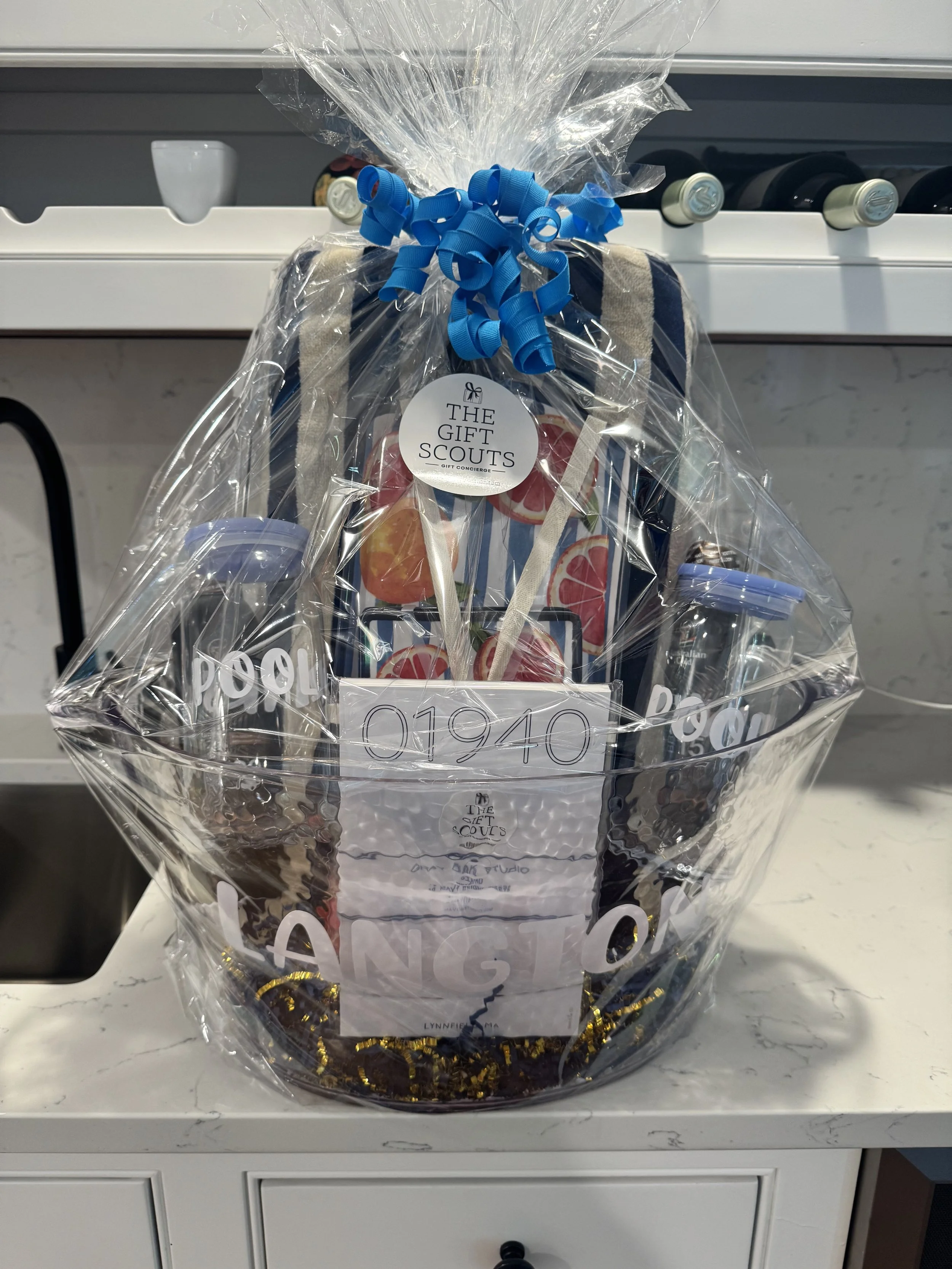 Gift basket wrapped in clear plastic with a blue curly ribbon on top, containing assorted snacks and items, sitting on a white surface in front of a white cabinet with bottles on top.