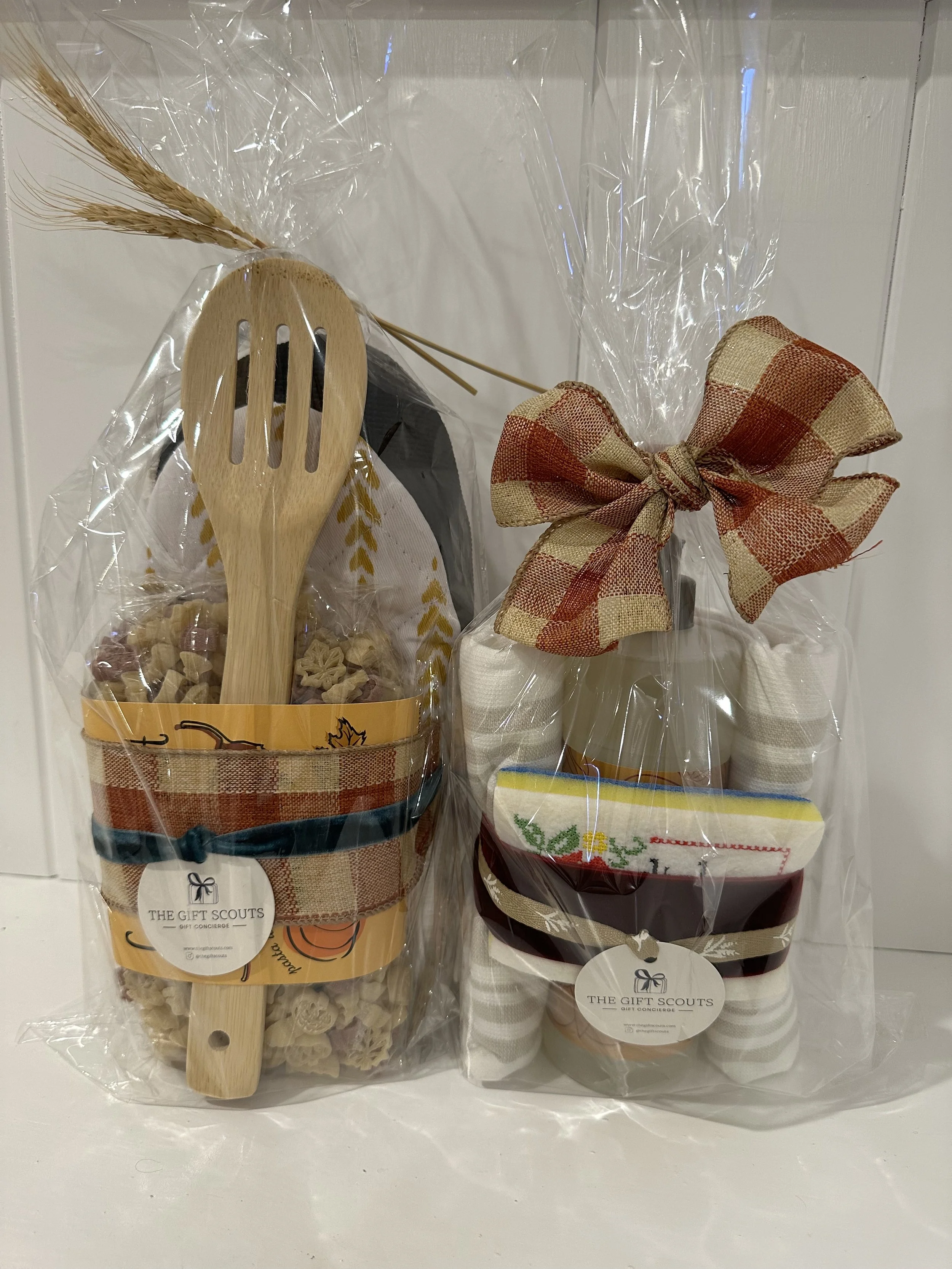Gift basket containing wooden spoon, pasta, and snack items in a clear plastic bag tied with a plaid ribbon, decorated with a tag reading 'The Gift Scouts'.