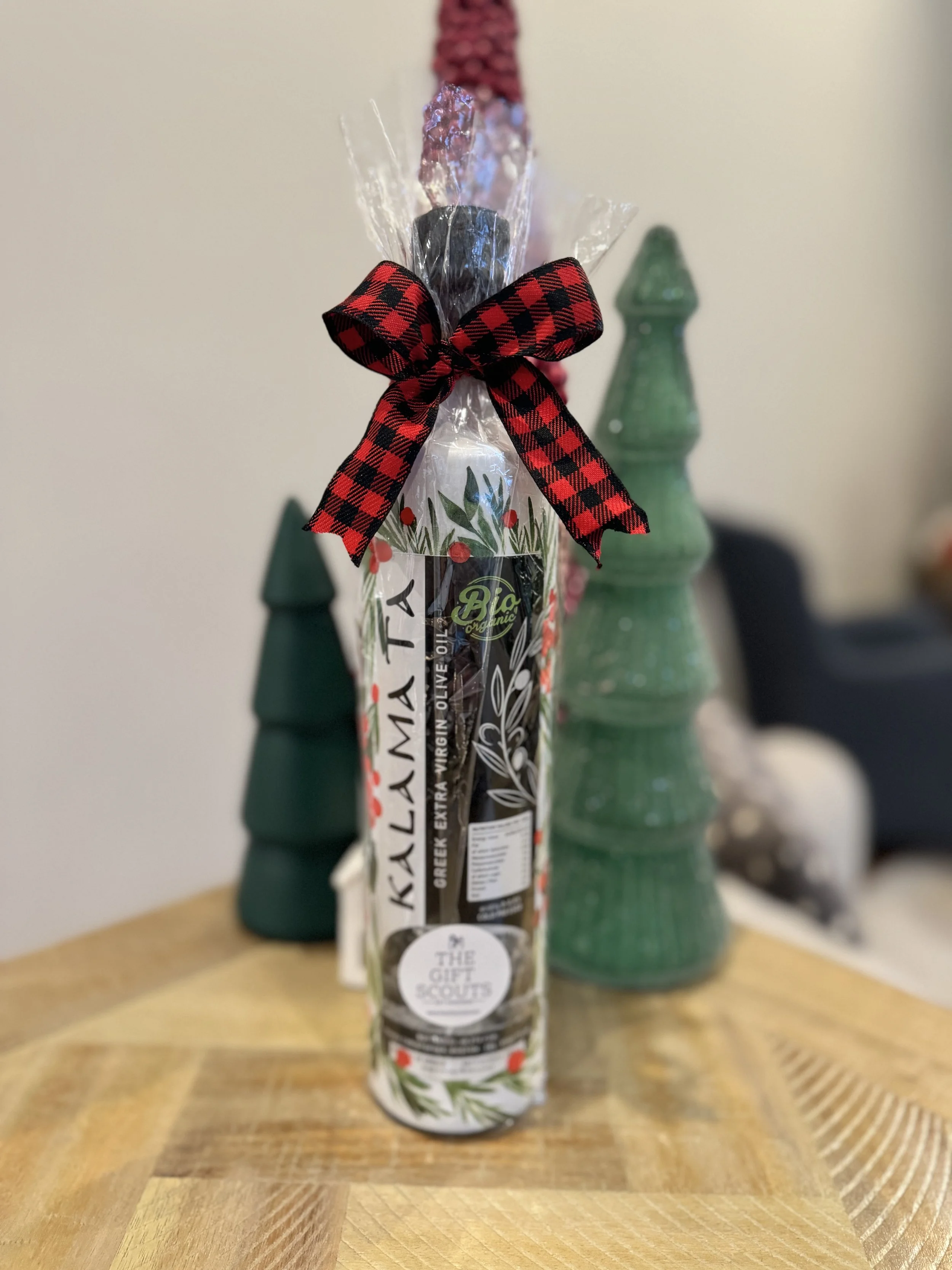 A bottle of Kalamata Greek extra virgin olive oil wrapped in clear plastic with a red and black checkered ribbon tied in a bow at the top, placed on a wooden surface with green ceramic Christmas tree decorations in the background.