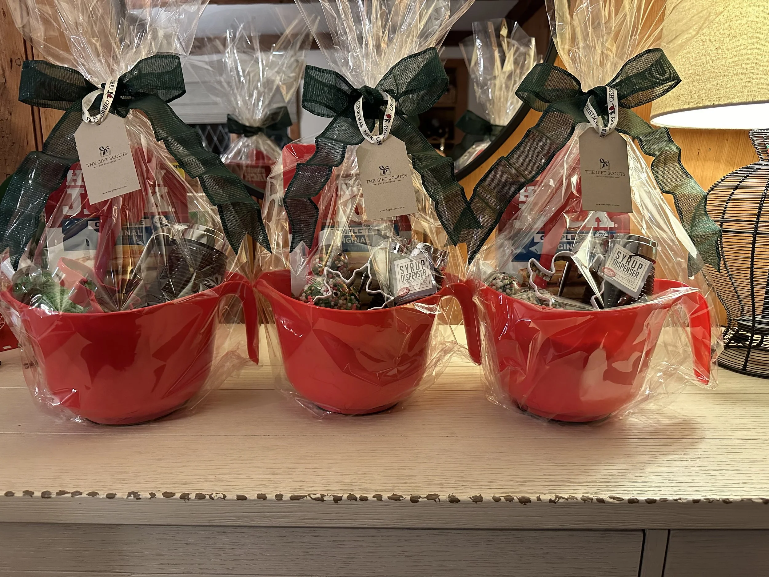 Three gift baskets wrapped in clear plastic with black ribbons, each placed in a red bowl, containing holiday-themed items like mugs and syrup, on a light-colored wooden surface.