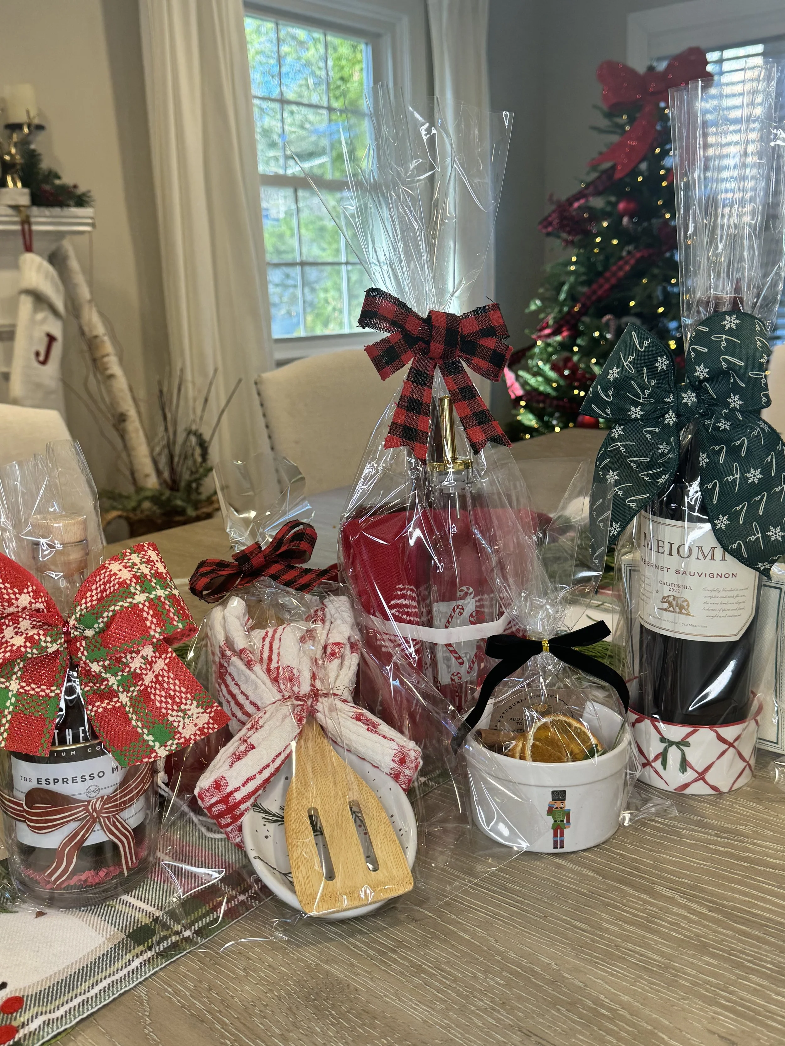 Holiday gift basket with wrapped bottles, cookies, and kitchen utensils, decorated with red and green bows, on a wooden table near a decorated Christmas tree.