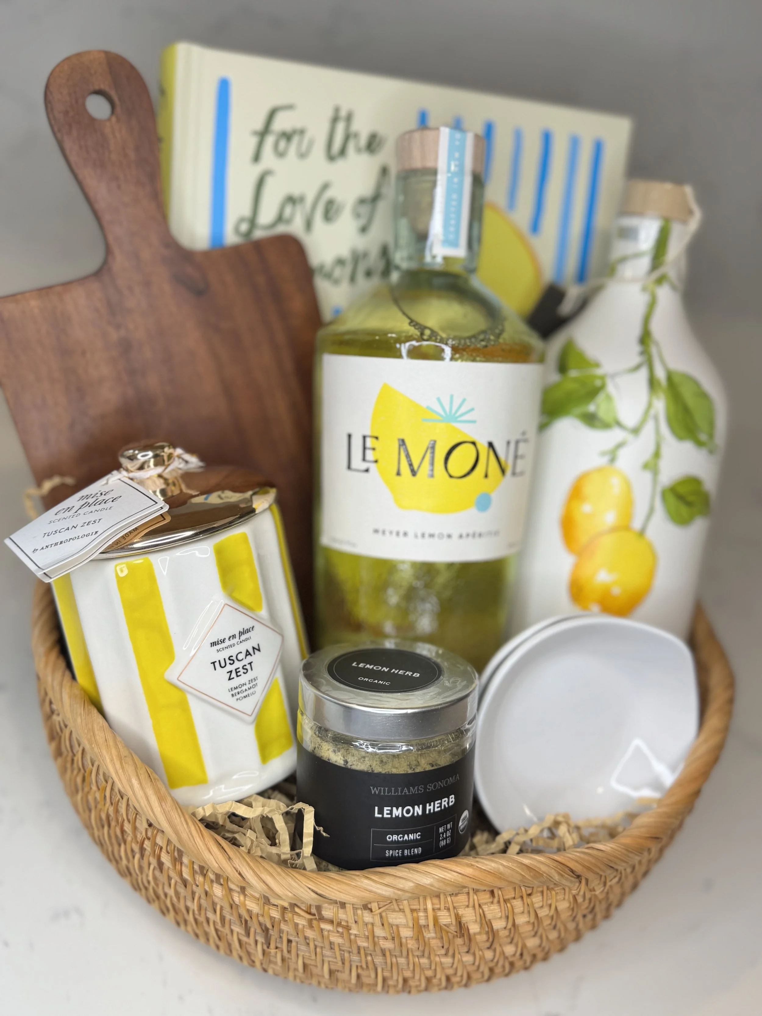A wicker basket containing lemon-themed items including a large bottle labeled 'Le Moné' with lemon illustrations, a jar of Williams Sonoma Lemon Herb spice, a container of Mise en Place Tuscan Zest, a white ceramic vase with lemon and green leaf des