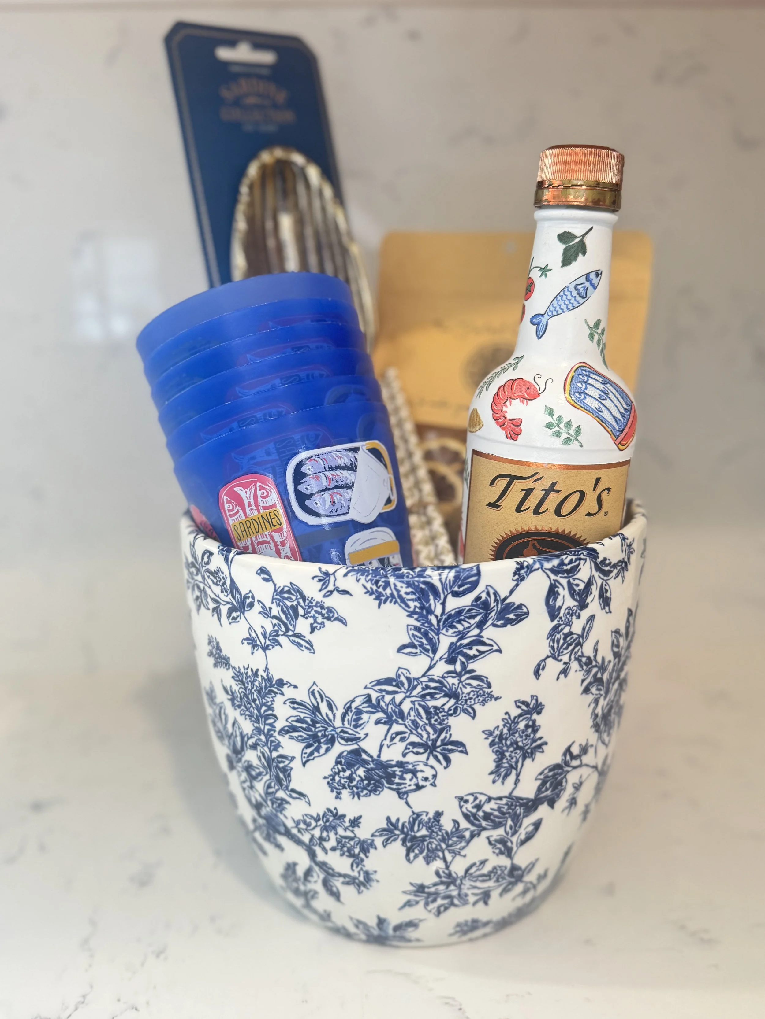 A white ceramic cup with blue floral pattern containing a bottle of Tito's vodka, a package of sardines, a wooden dish, and a stack of blue plastic cups with sardine and fish stickers.