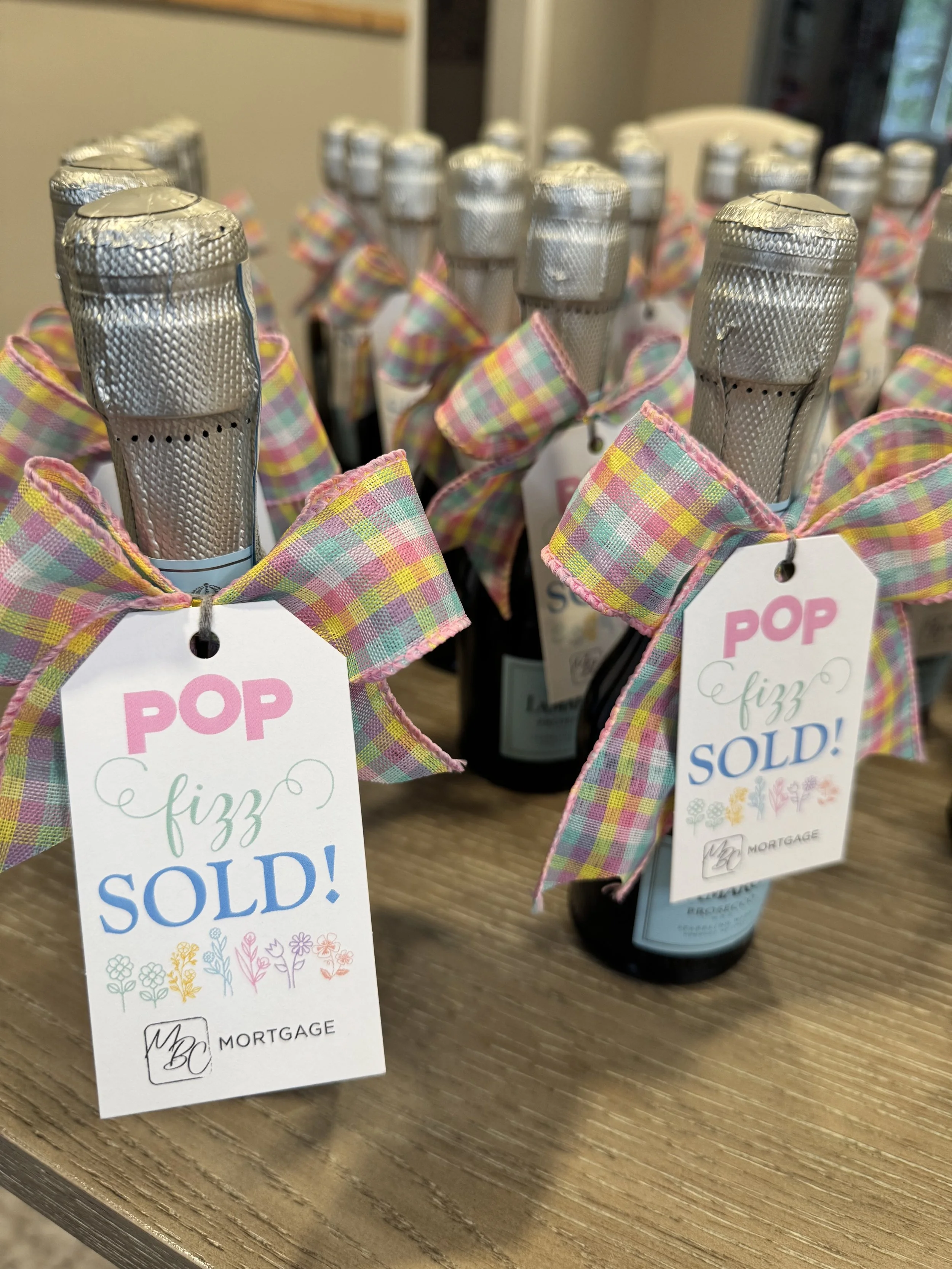 Multiple small bottles with silver foil caps and colorful checkered ribbons tied around their necks, each with a tag that reads 'POP fizz SOLD!' and a logo for MBC Mortgage, arranged on a wooden surface.