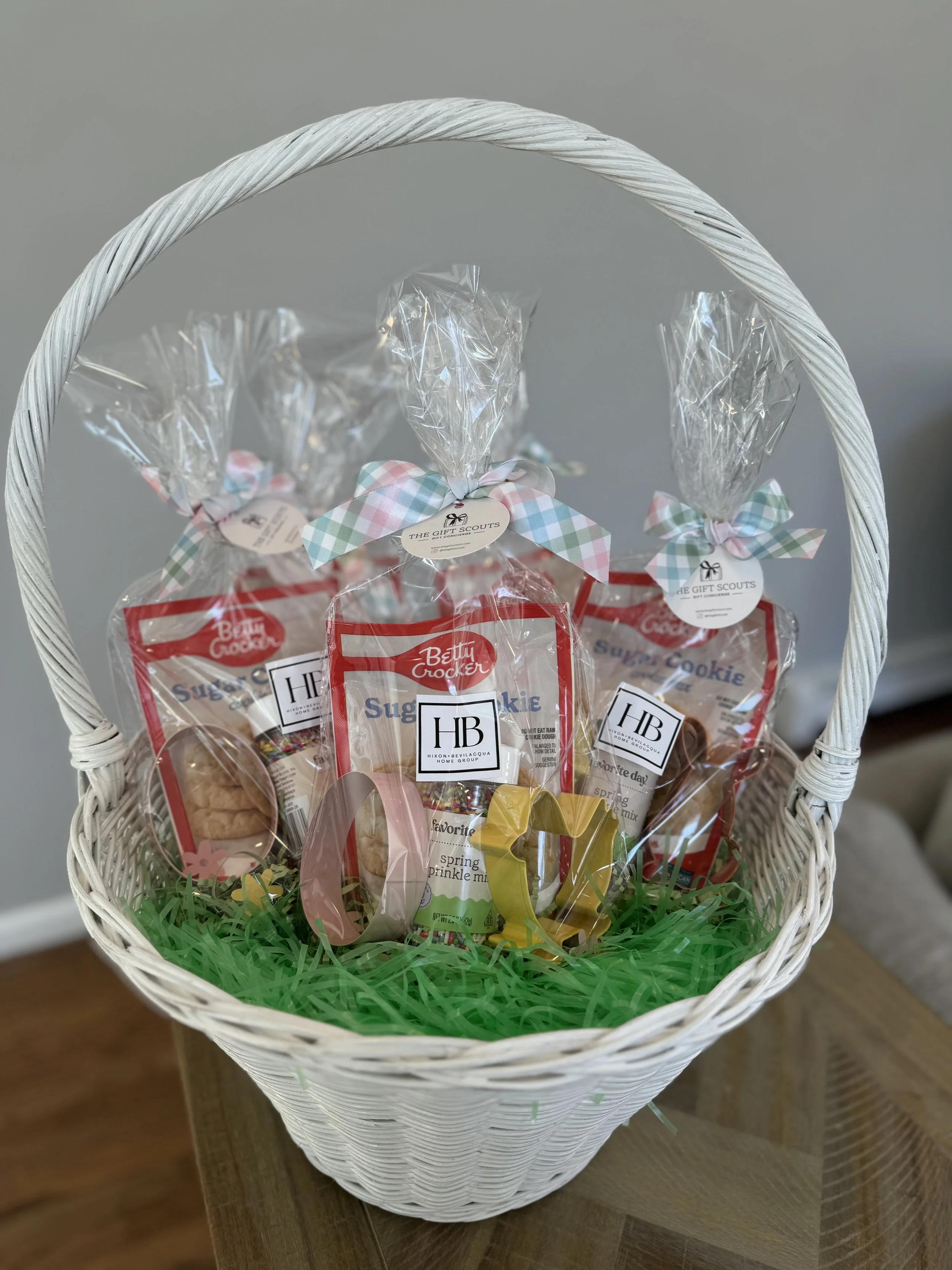 Easter gift basket with green shredded paper, containing Betty Crocker sugar cookie kits, decorated with pastel ribbons and small decorative yellow, pink, and gold balloons.