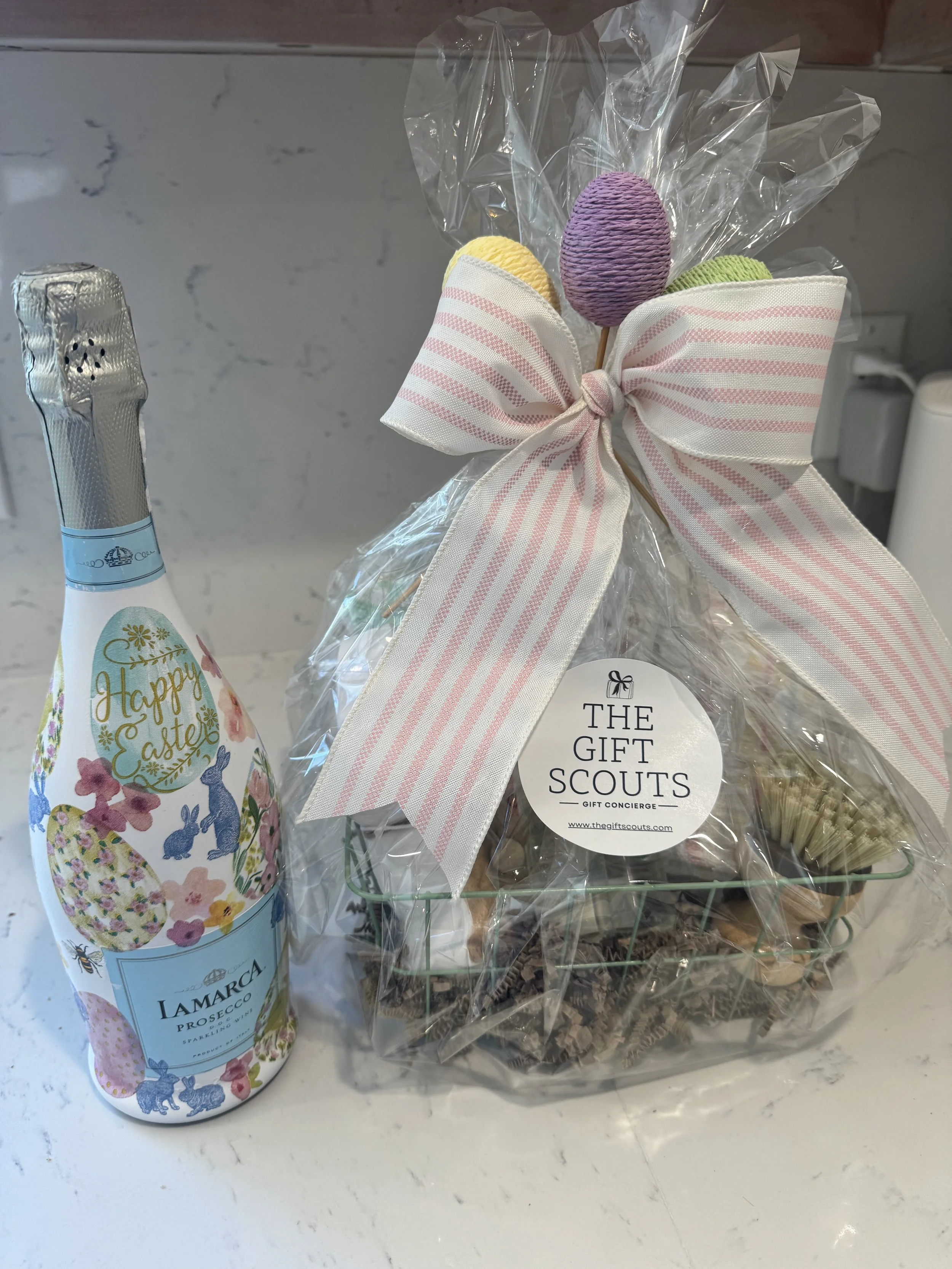 Decorative Easter gift basket wrapped in clear cellophane with pastel-colored candy eggs and a pink and white striped bow, along with a bottle of Lamarca Prosecco with Easter-themed label featuring bunnies and eggs.