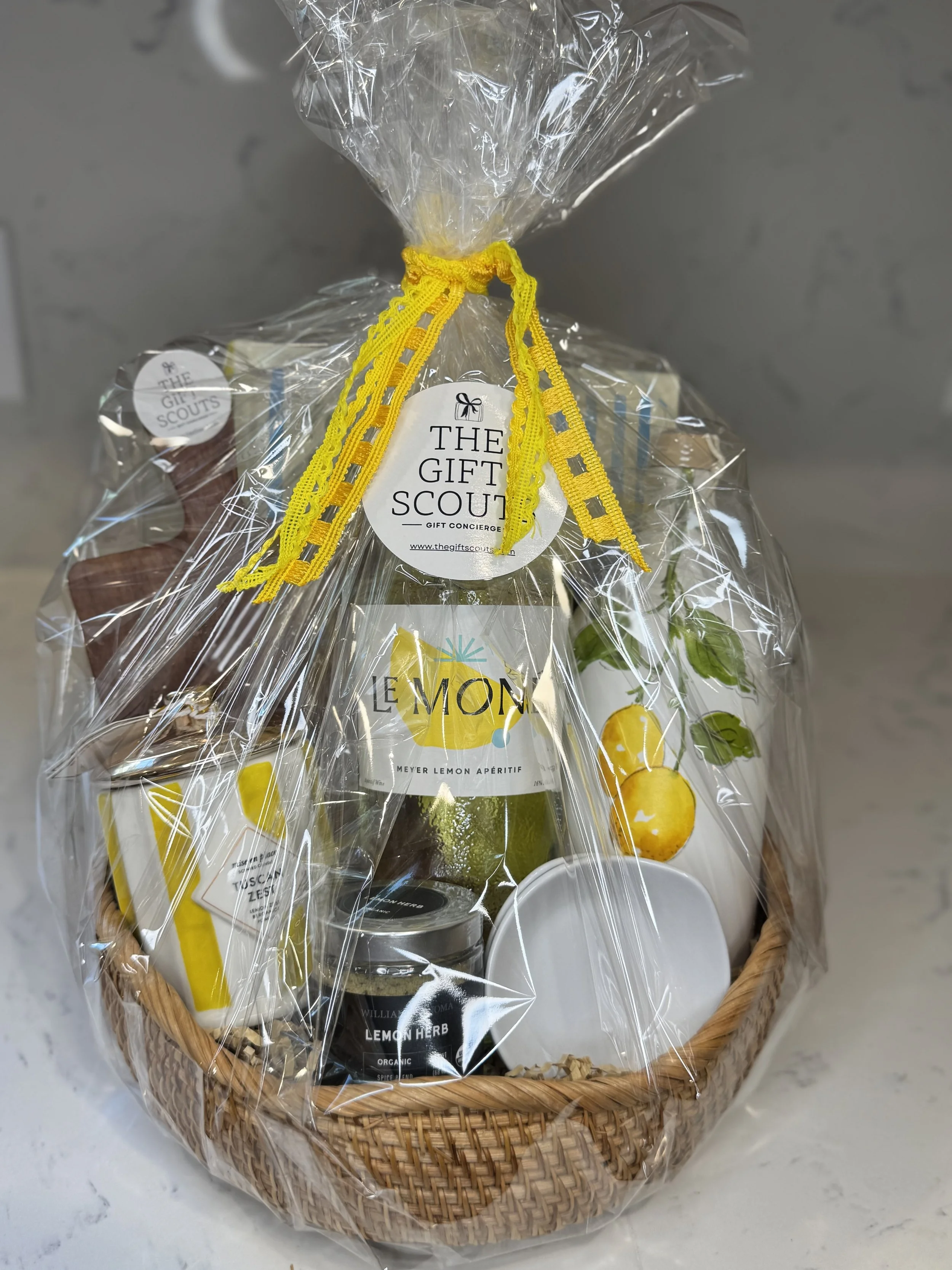 Gift basket wrapped in clear cellophane with a yellow ribbon. Contains lemon-themed items including a bottle, jar, and mug, along with a white ceramic dish.