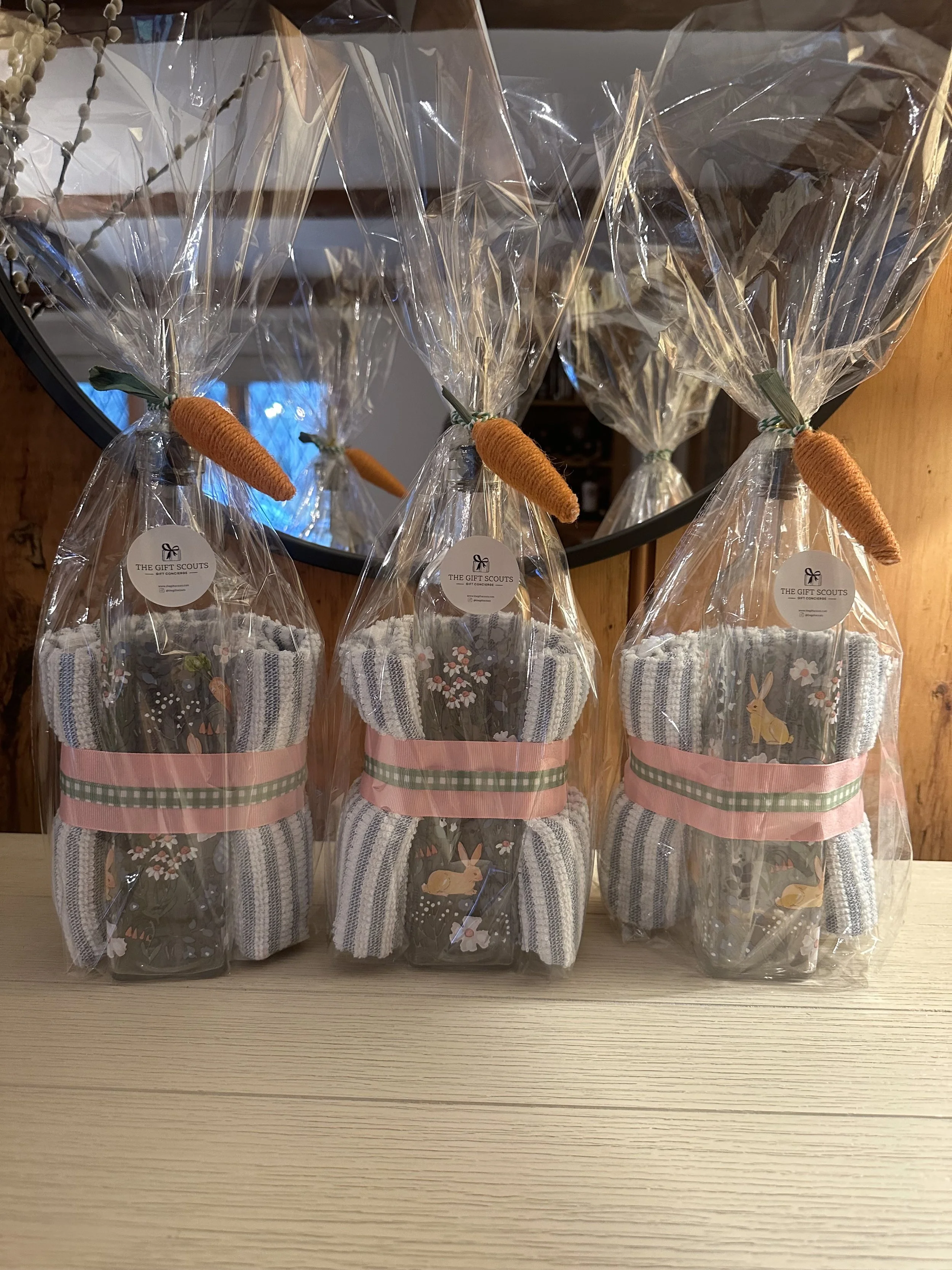 Three gift sets wrapped in clear plastic with decorative carrots on top, containing patterned socks and ribbons, placed on a wooden surface in front of a mirror.