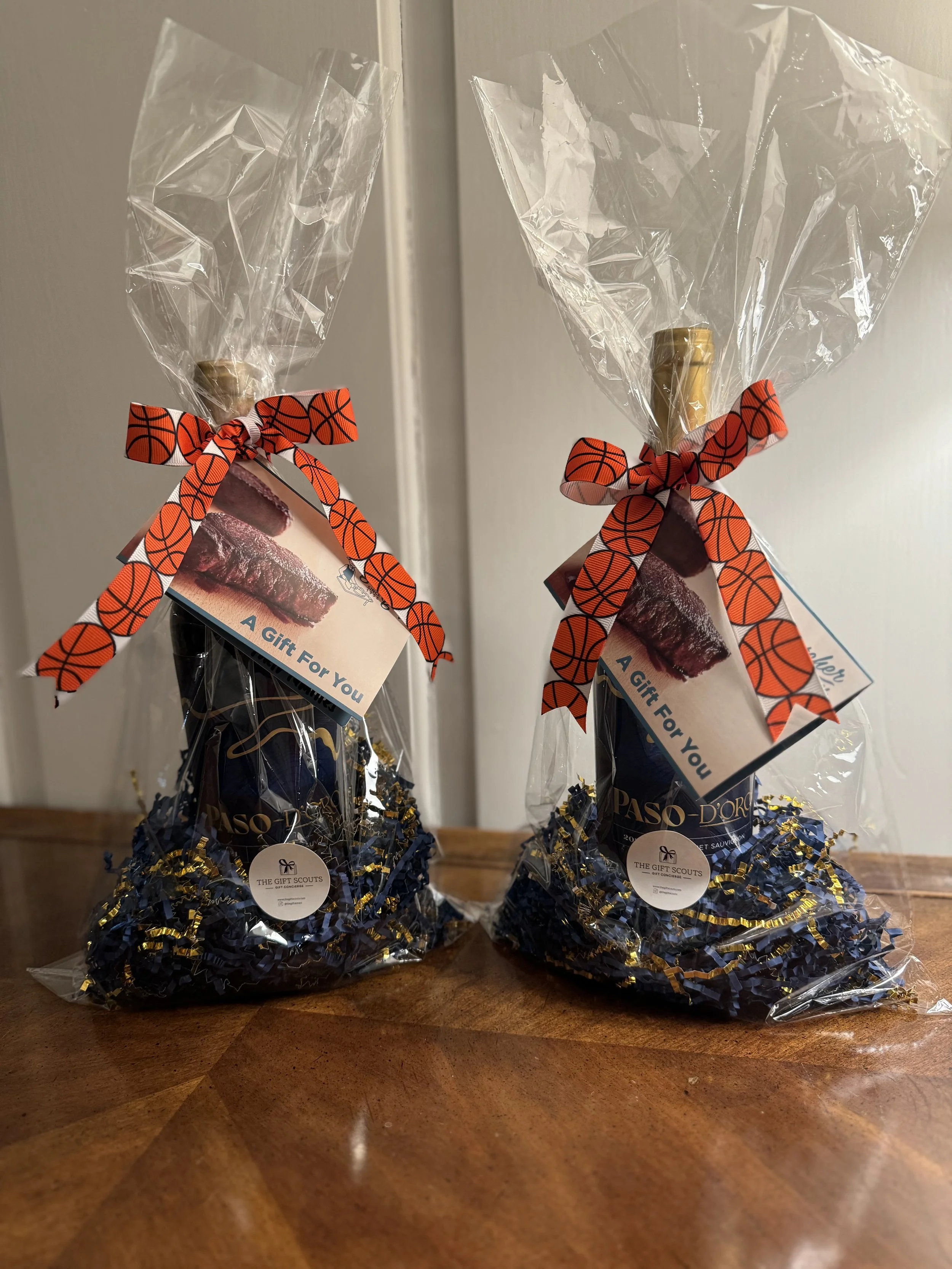 Two gift packages wrapped in clear plastic with orange ribbon featuring a basketball pattern, each containing chocolates and other treats on a wooden surface.