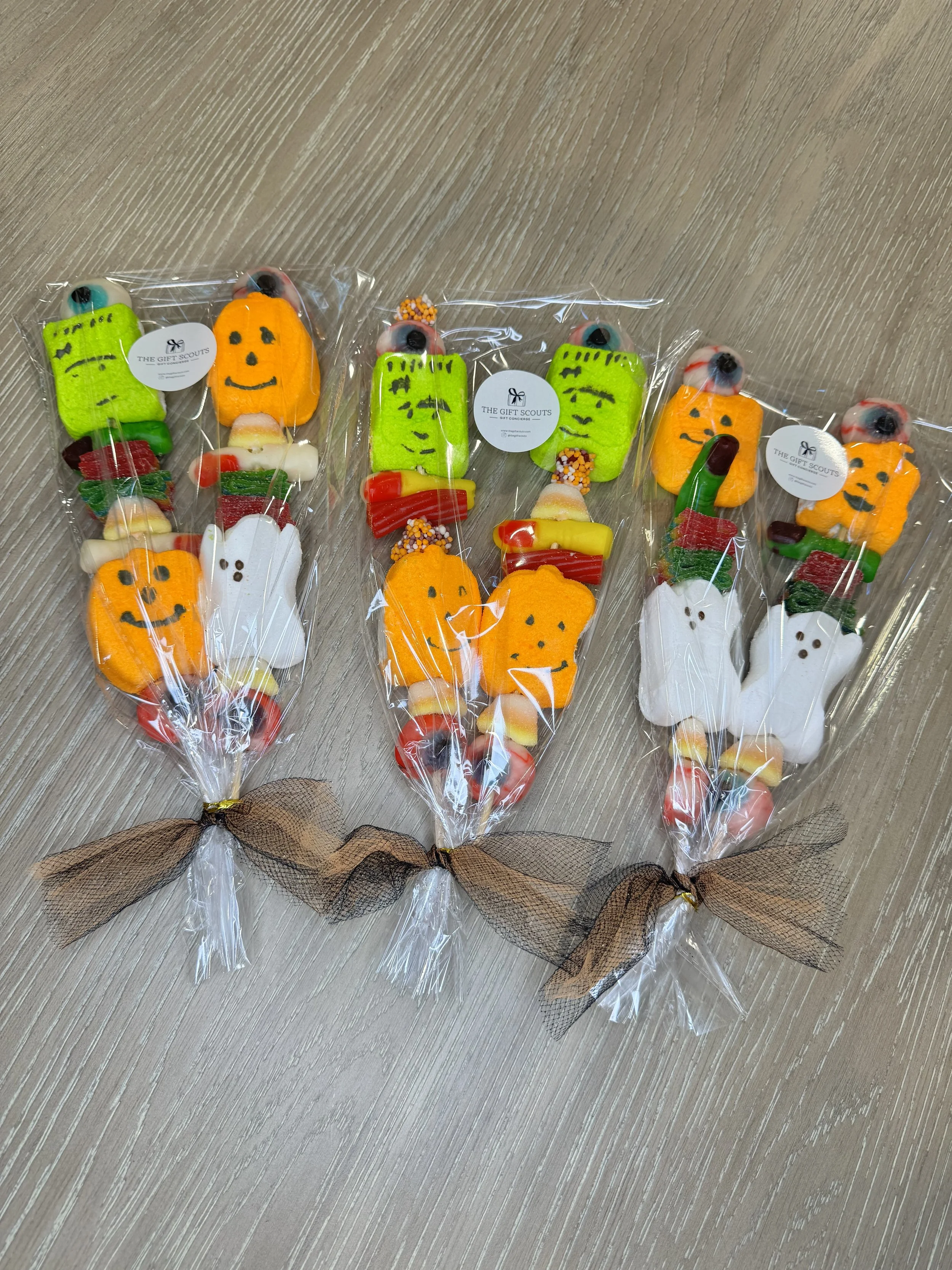 Three Halloween-themed cookie lollipops wrapped in plastic with brown ribbon, shaped like ghosts, pumpkins, and monsters, on a wooden surface.