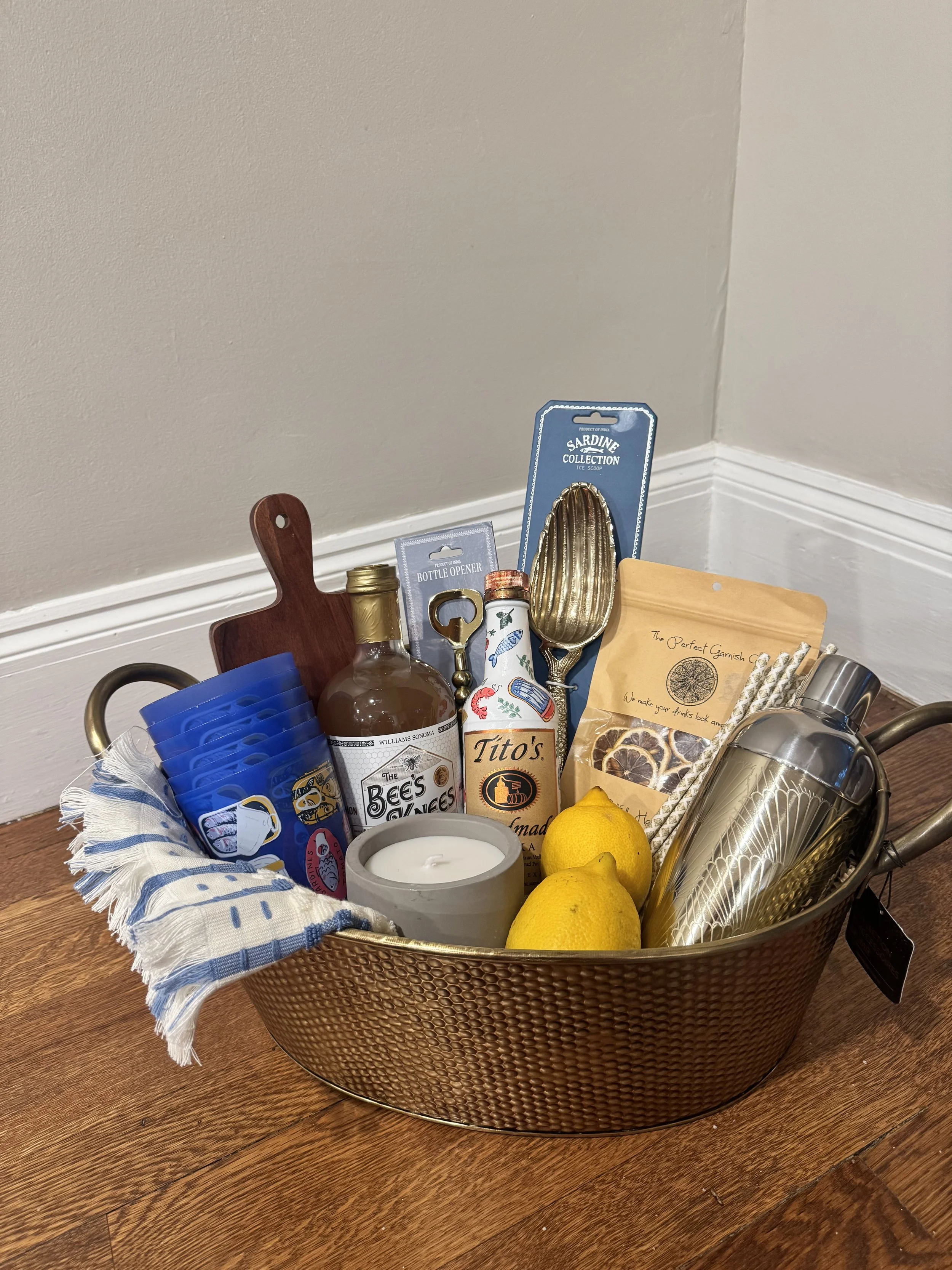 A metal basket on a wooden surface containing various items including two lemons, a white candle, a silver cocktail shaker, a bottle of sauce labeled 'The Bee's Knees,' a pack of gourmet lemon or orange slices, a blue face mask, a wooden cutting boar