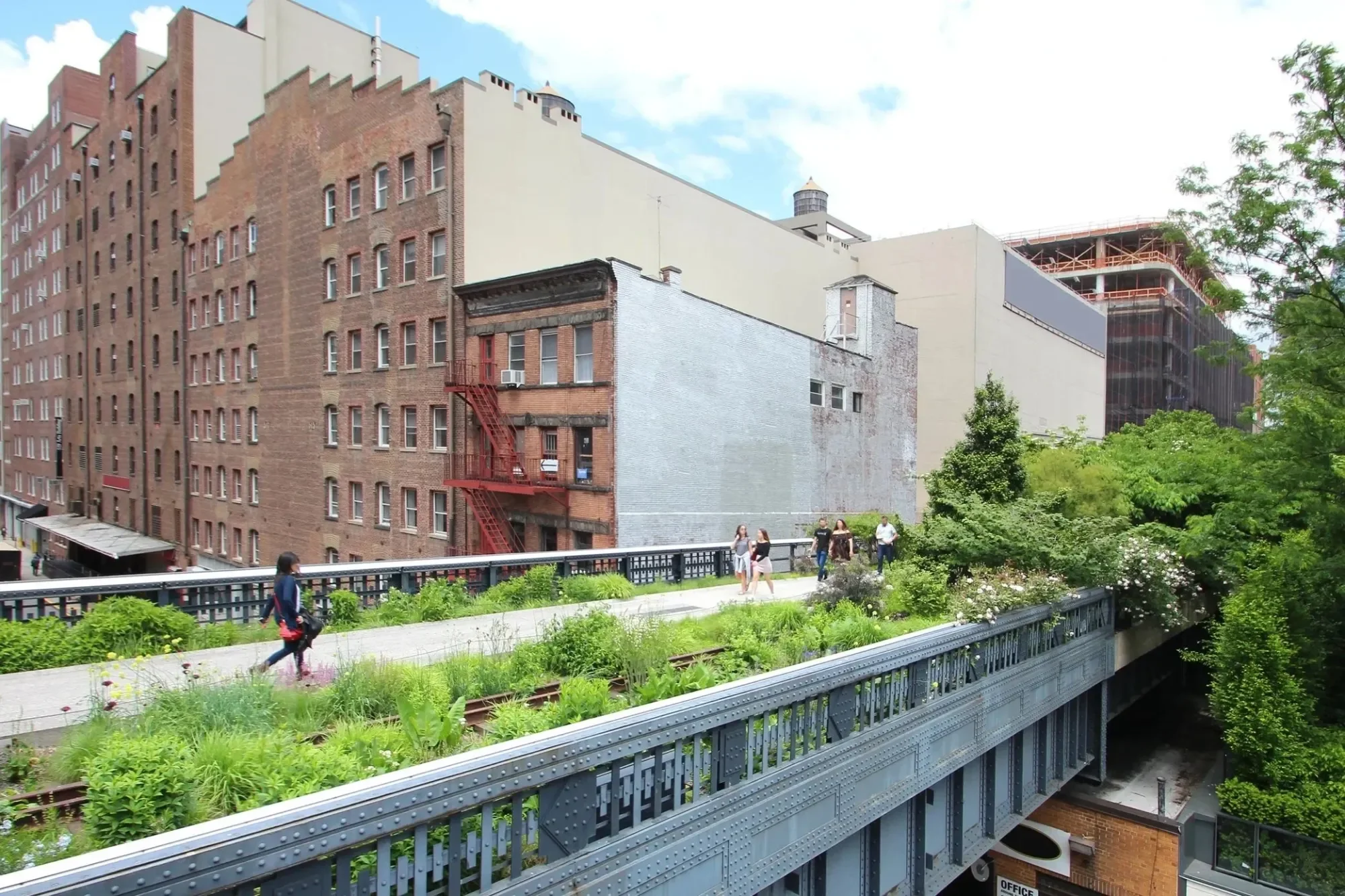 People walk along a green, elevated park lined with plants and pass by old brick and green buildings in the city.