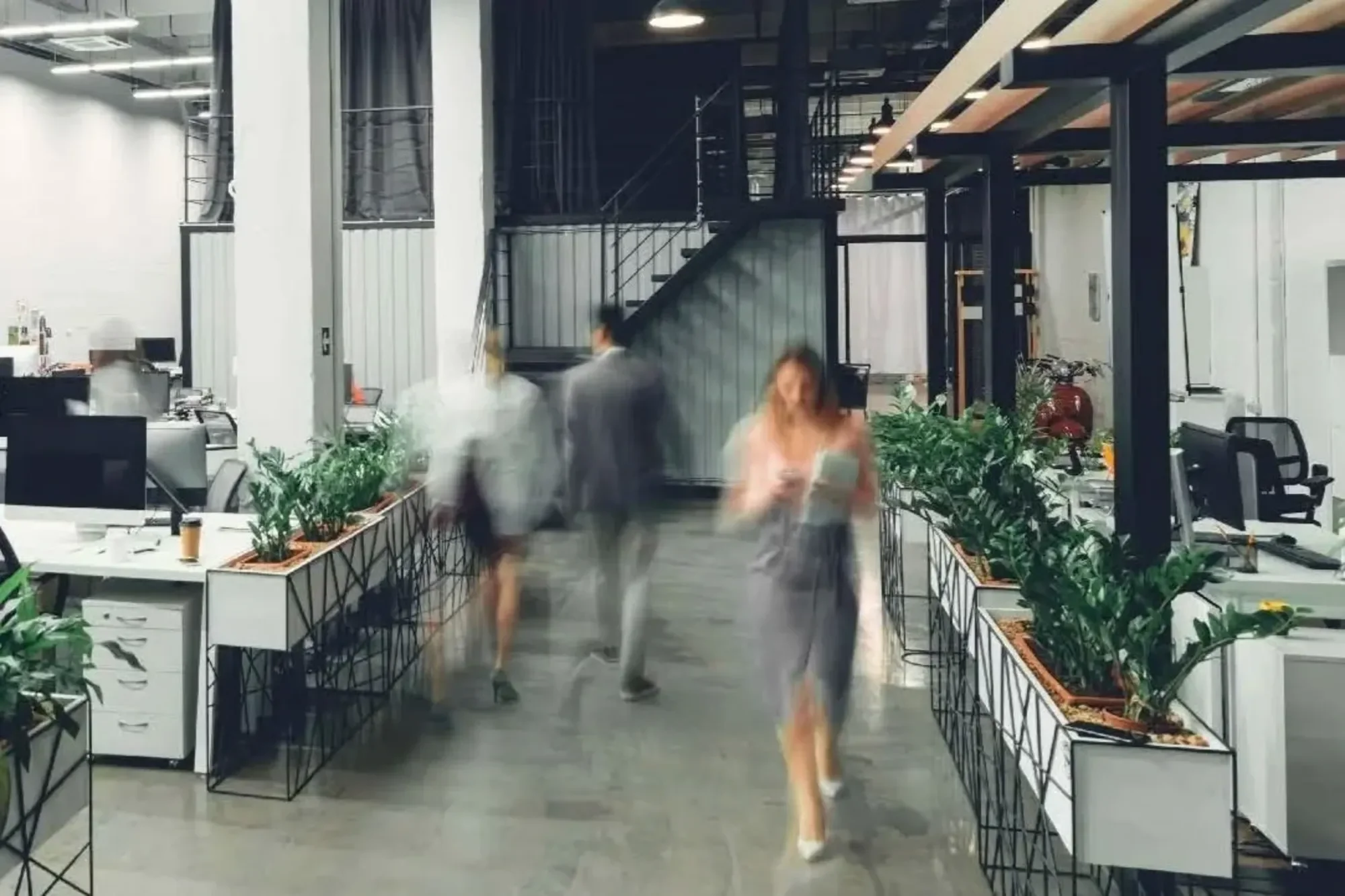 People walk through a modern green building office with desks, plants, and industrial-style decor; some appear blurred in motion.