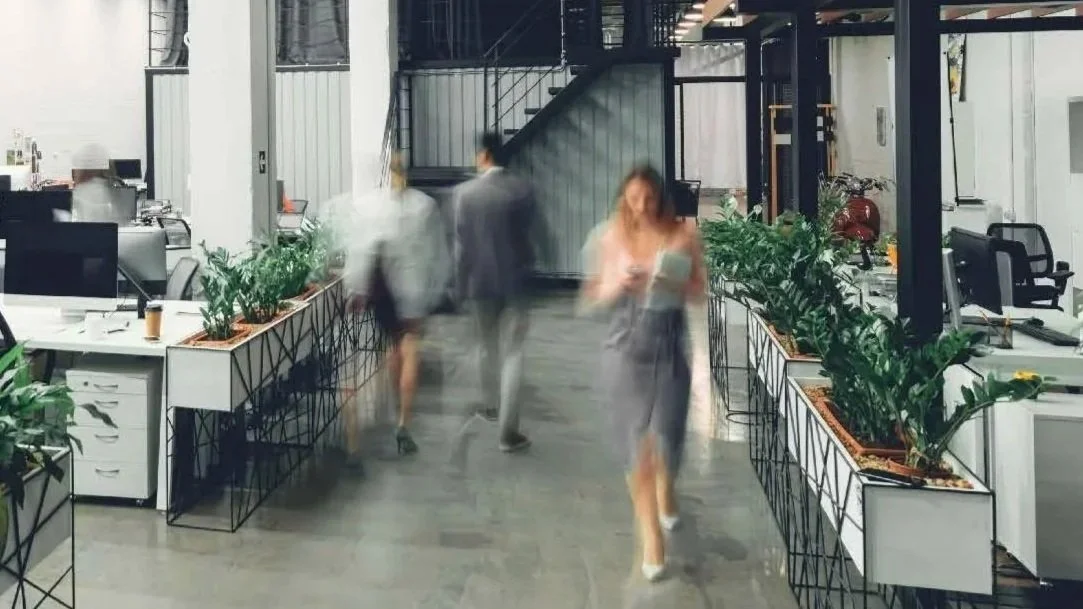 Blurry motion photo of a woman walking quickly through a modern indoor walkway with potted green plants and industrial-style architecture.