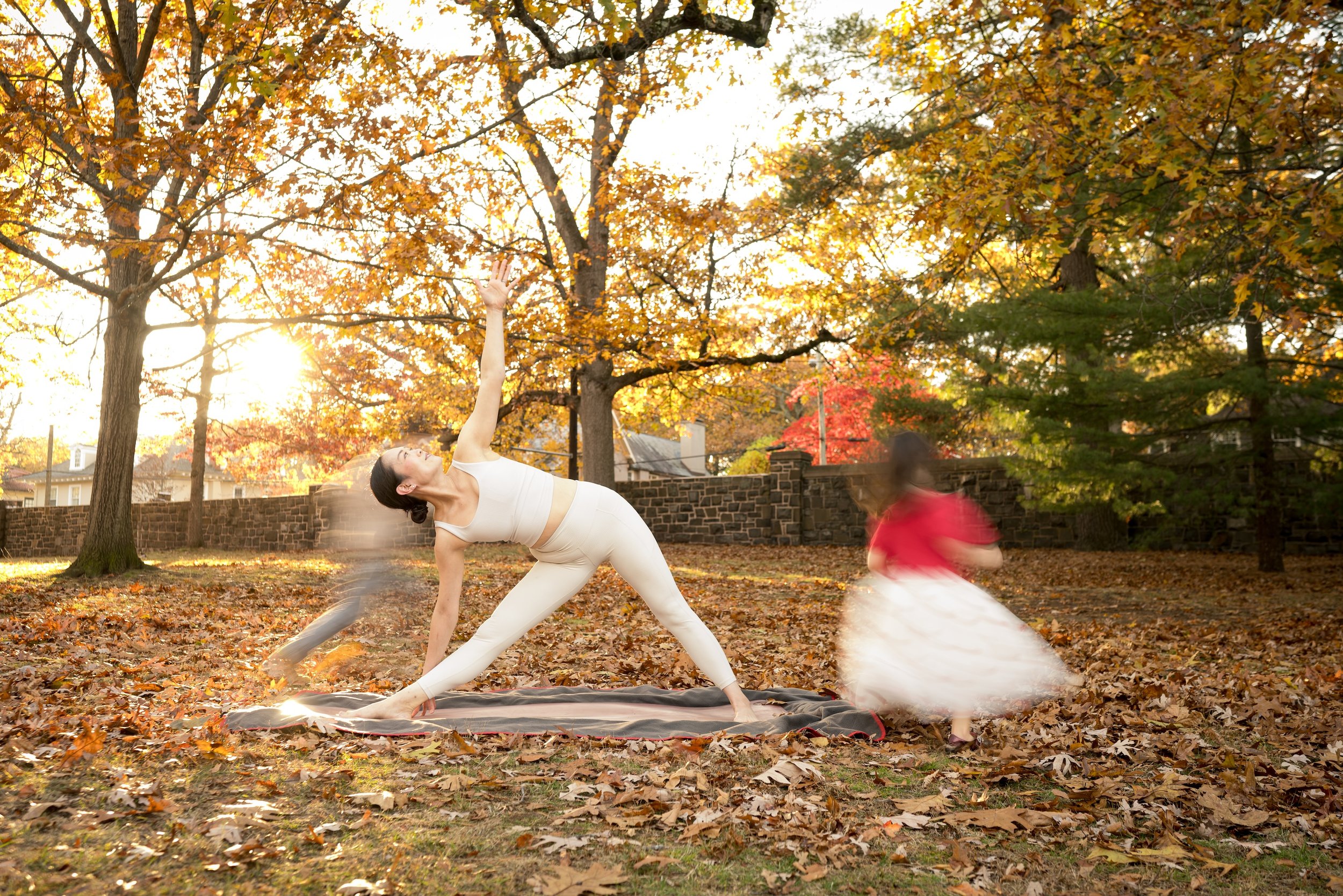 A woman practicing yoga outdoors on a fall day with autumn trees and fallen leaves, accompanied by a small girl in a red dress. The woman is in a side plank pose with her arms raised, and the girl is spinning around, blurred by movement.