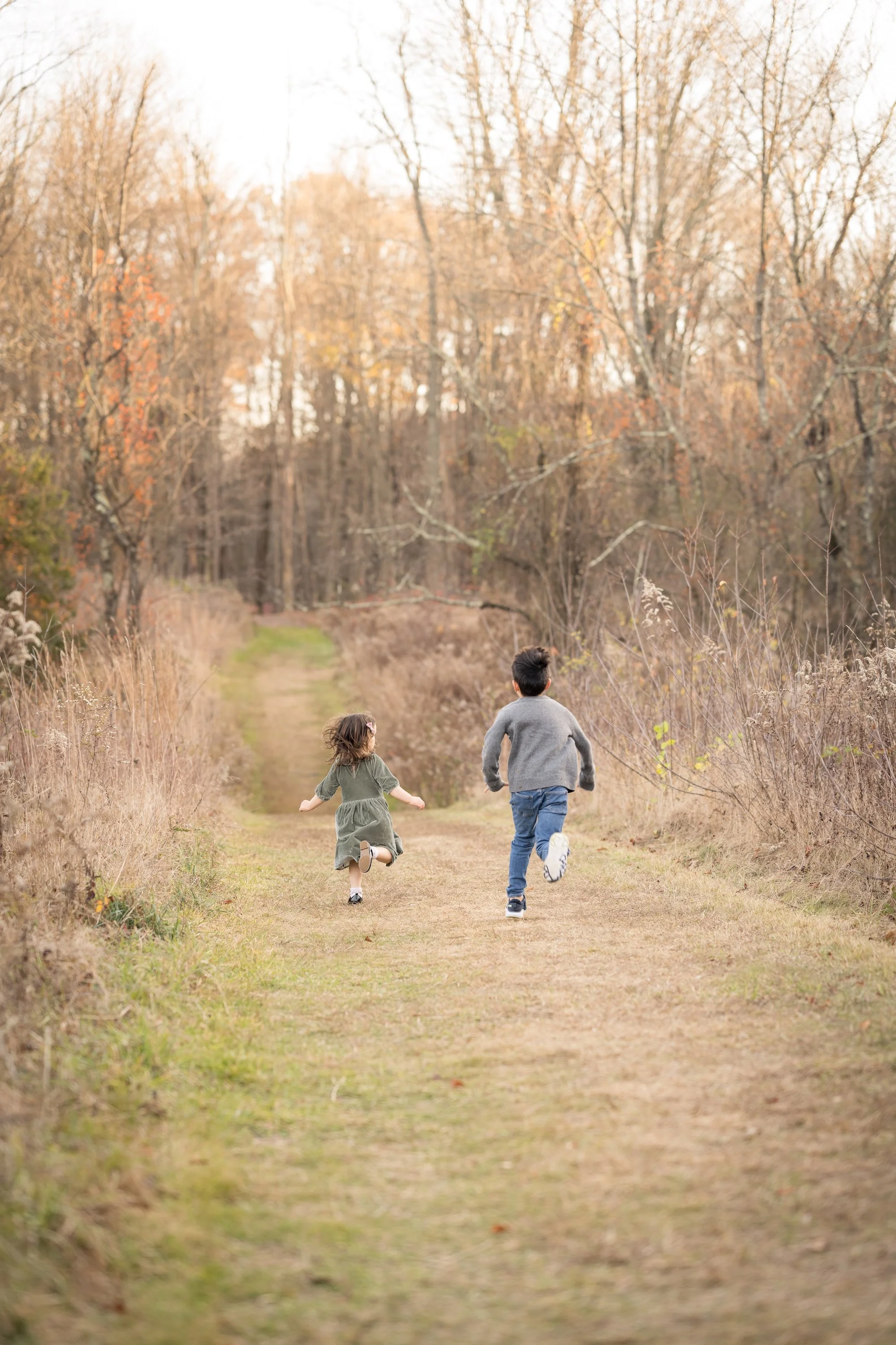 Two children running along a dirt path in a fall wooded landscape with leafless trees.