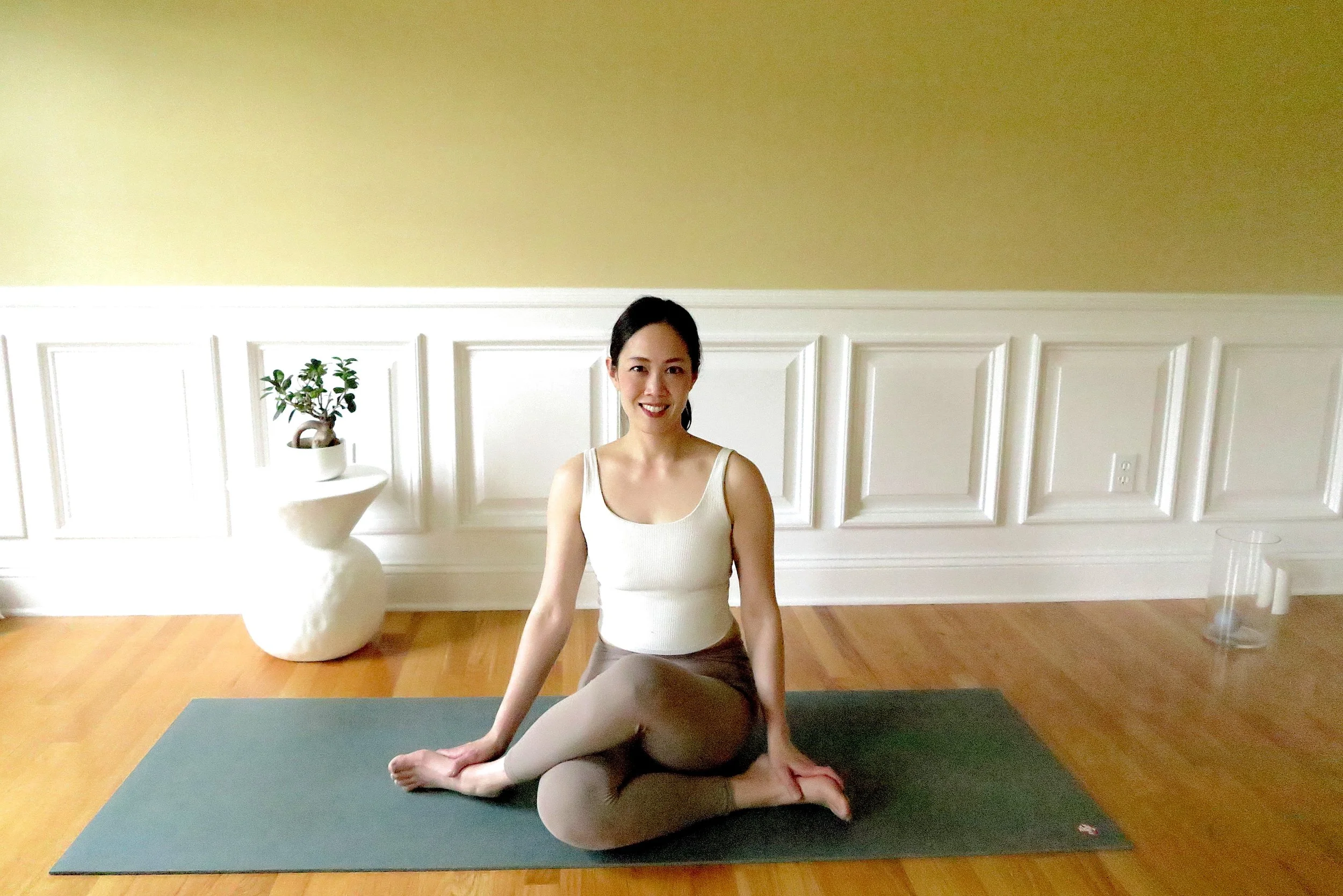 A woman practicing yoga seated on a mat in a spacious room with white wainscoting and a beige wall. She is smiling, wearing a white tank top and beige leggings, with a small potted plant on a white pedestal nearby.