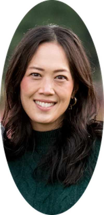 A woman with dark wavy hair smiling outdoors with a blurred green background.