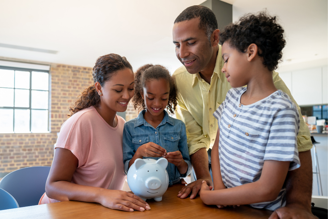 Family with two children smiling as a child puts a coin into a piggy bank on a table at home.